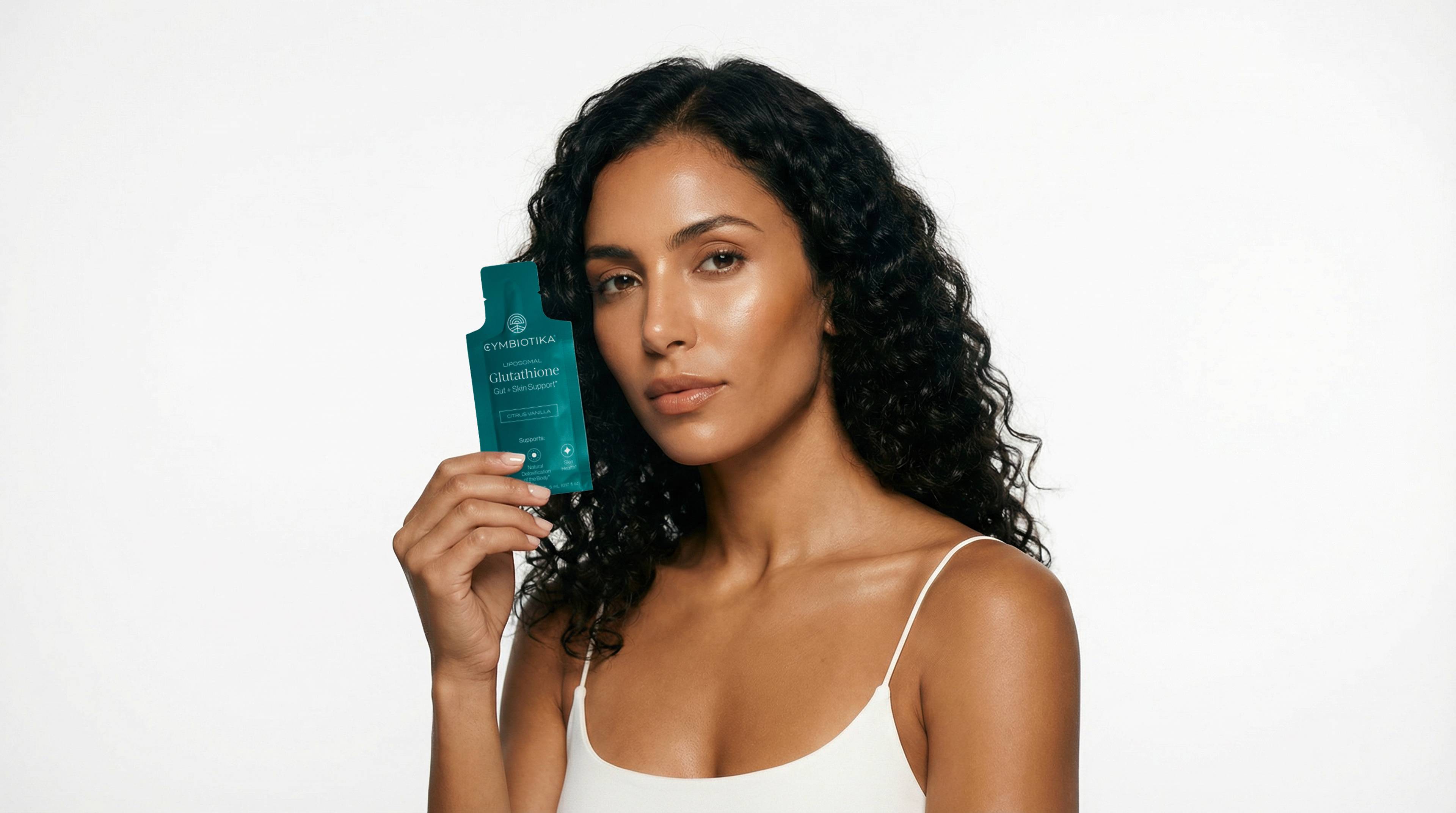 Woman in a white camisole holds a Cymbiotika Liposomal Glutathione packet beside her face.