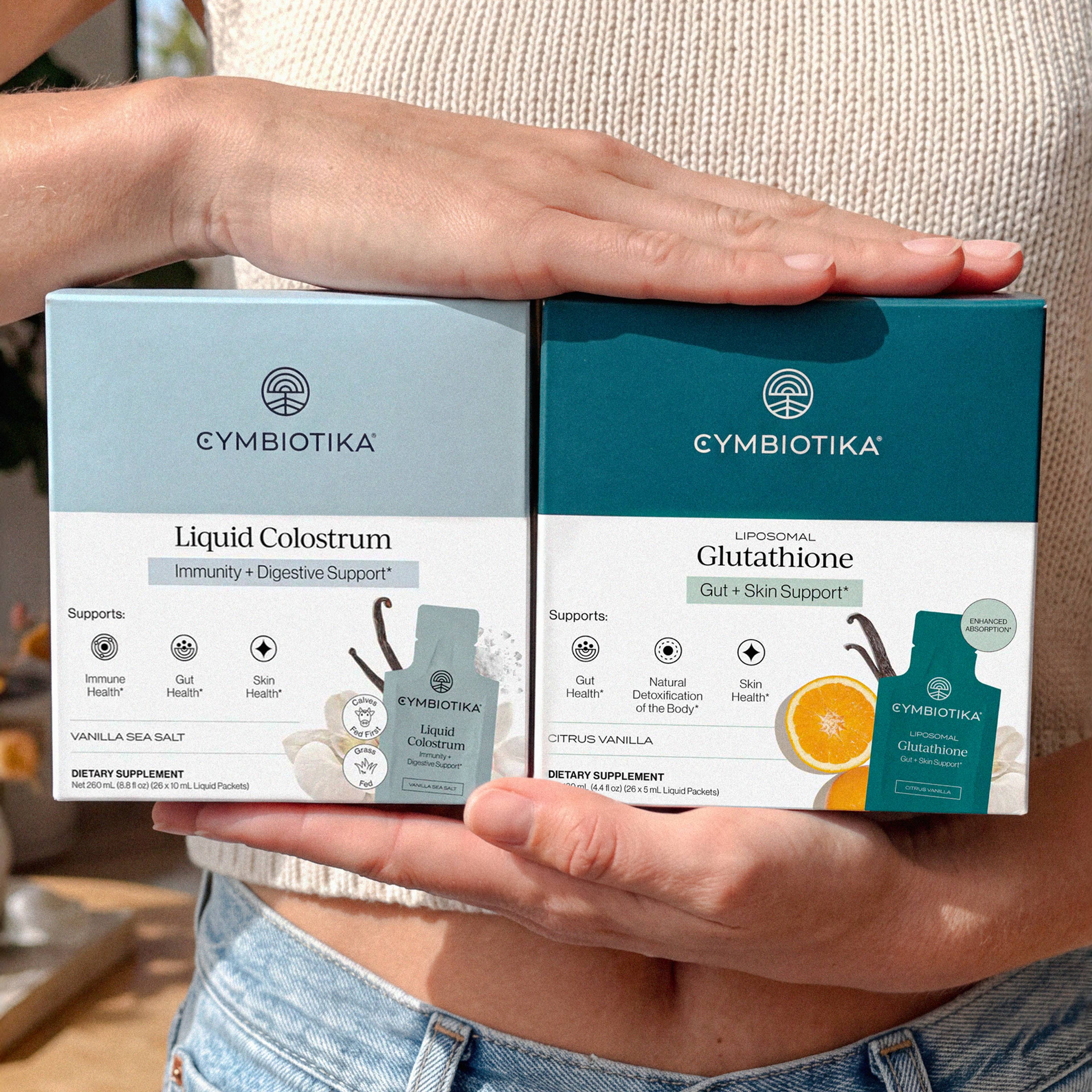 A person holds boxes of Cymbiotika Liquid Colostrum and Liposomal Glutathione.