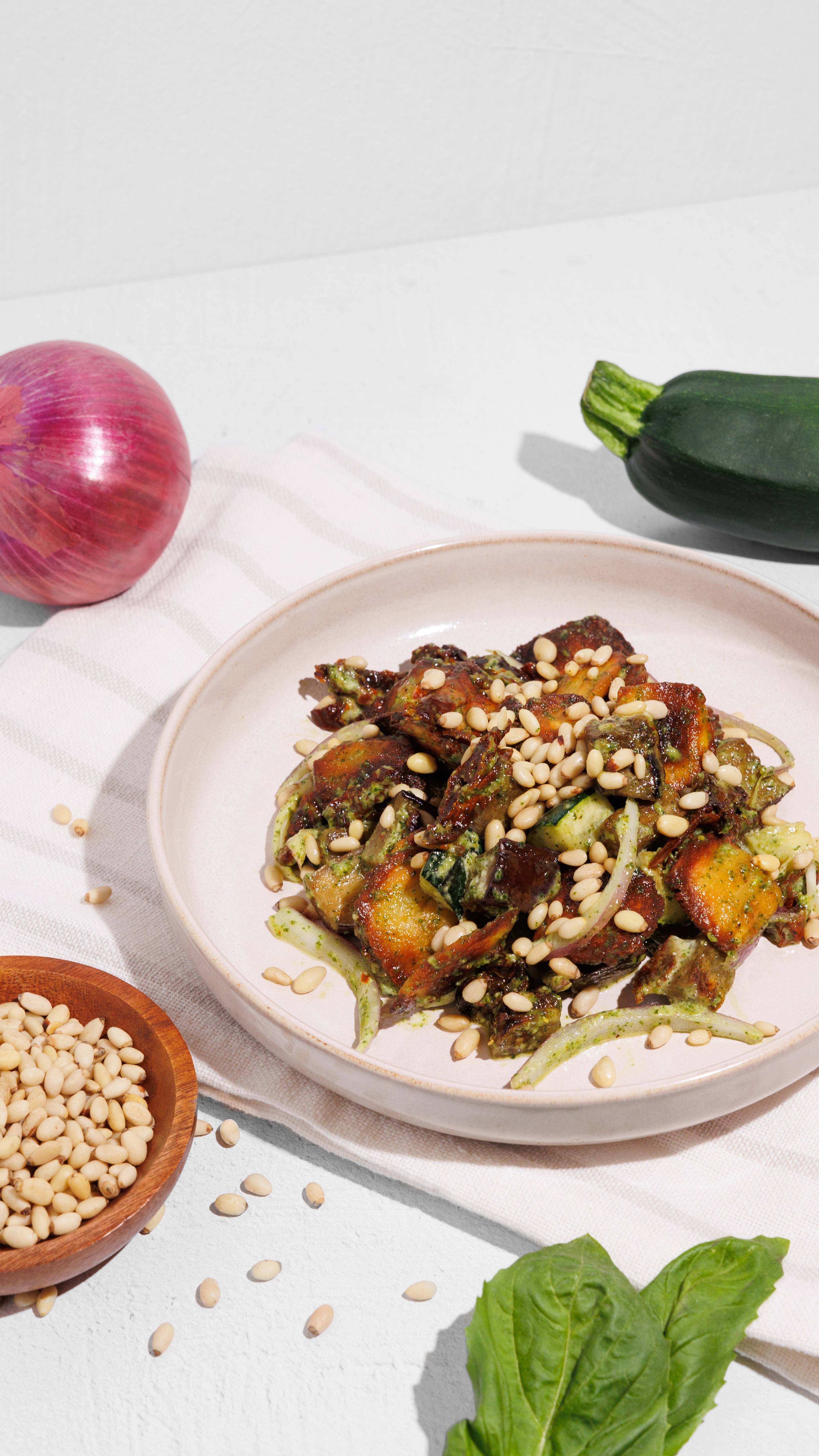 Plate of crispy gnocchi salad with pesto, zucchini, onion, and pine nuts, surrounded by fresh ingredients.