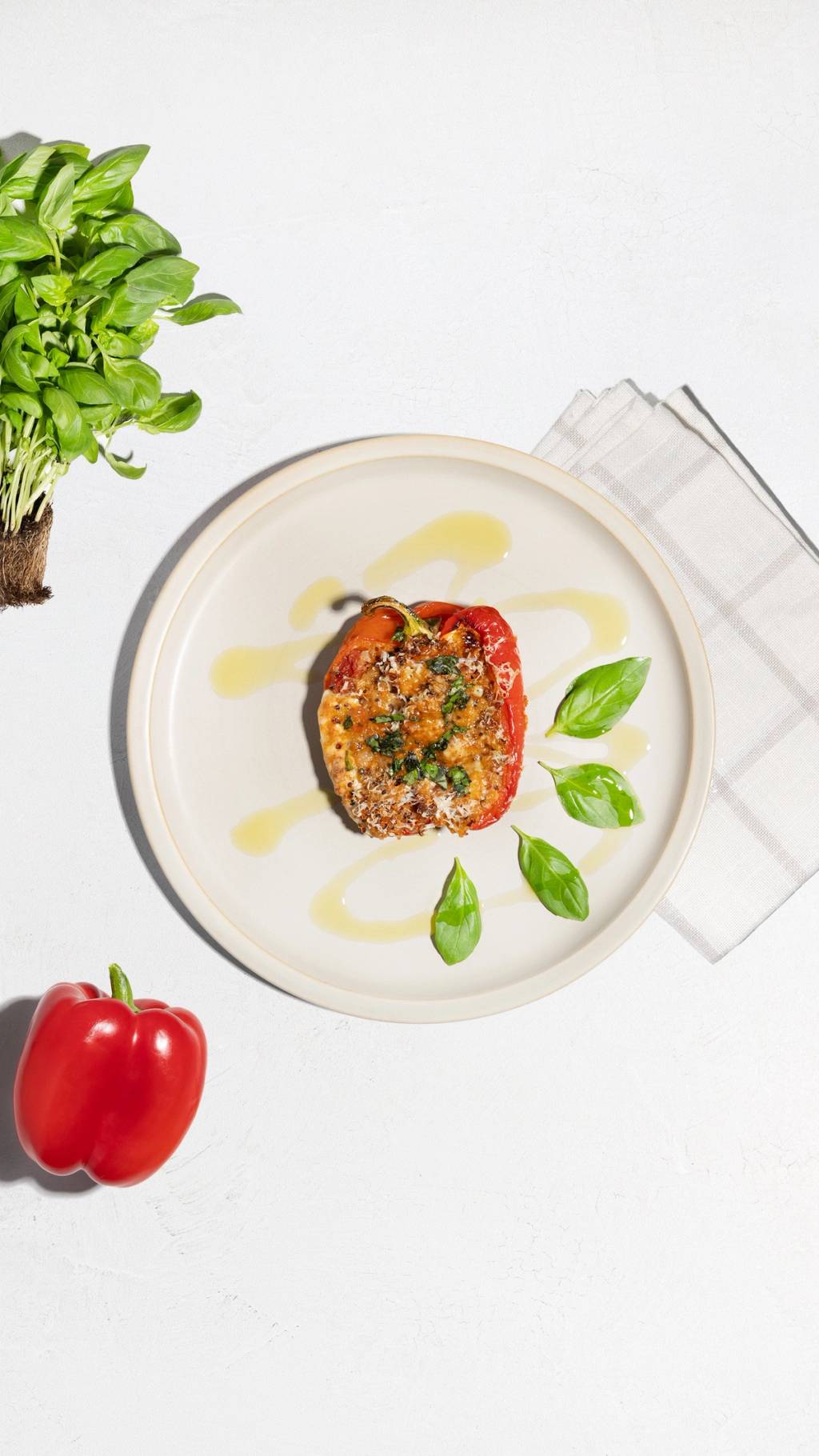 Stuffed red bell pepper with basil and oil on a plate, beside fresh basil and a whole pepper.