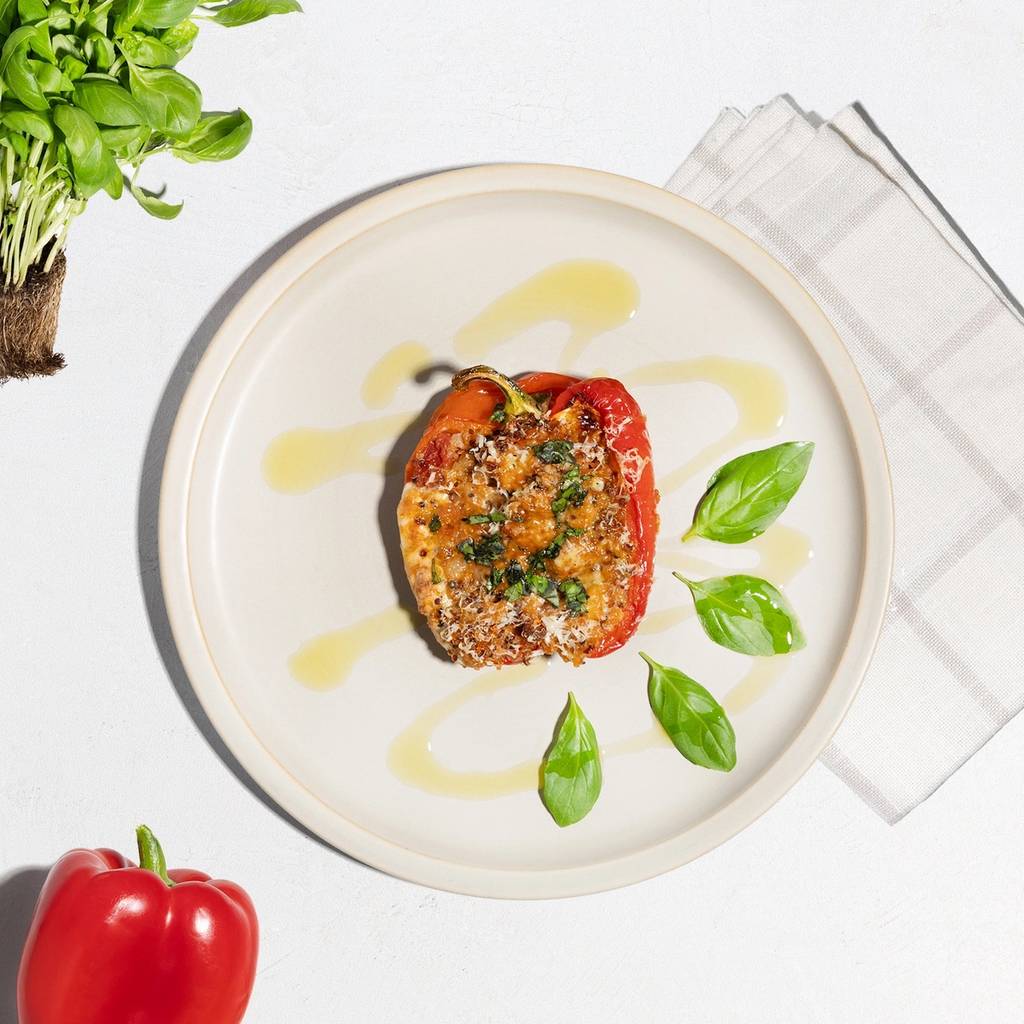 Stuffed red bell pepper with basil and oil on a plate, beside fresh basil and a whole pepper.