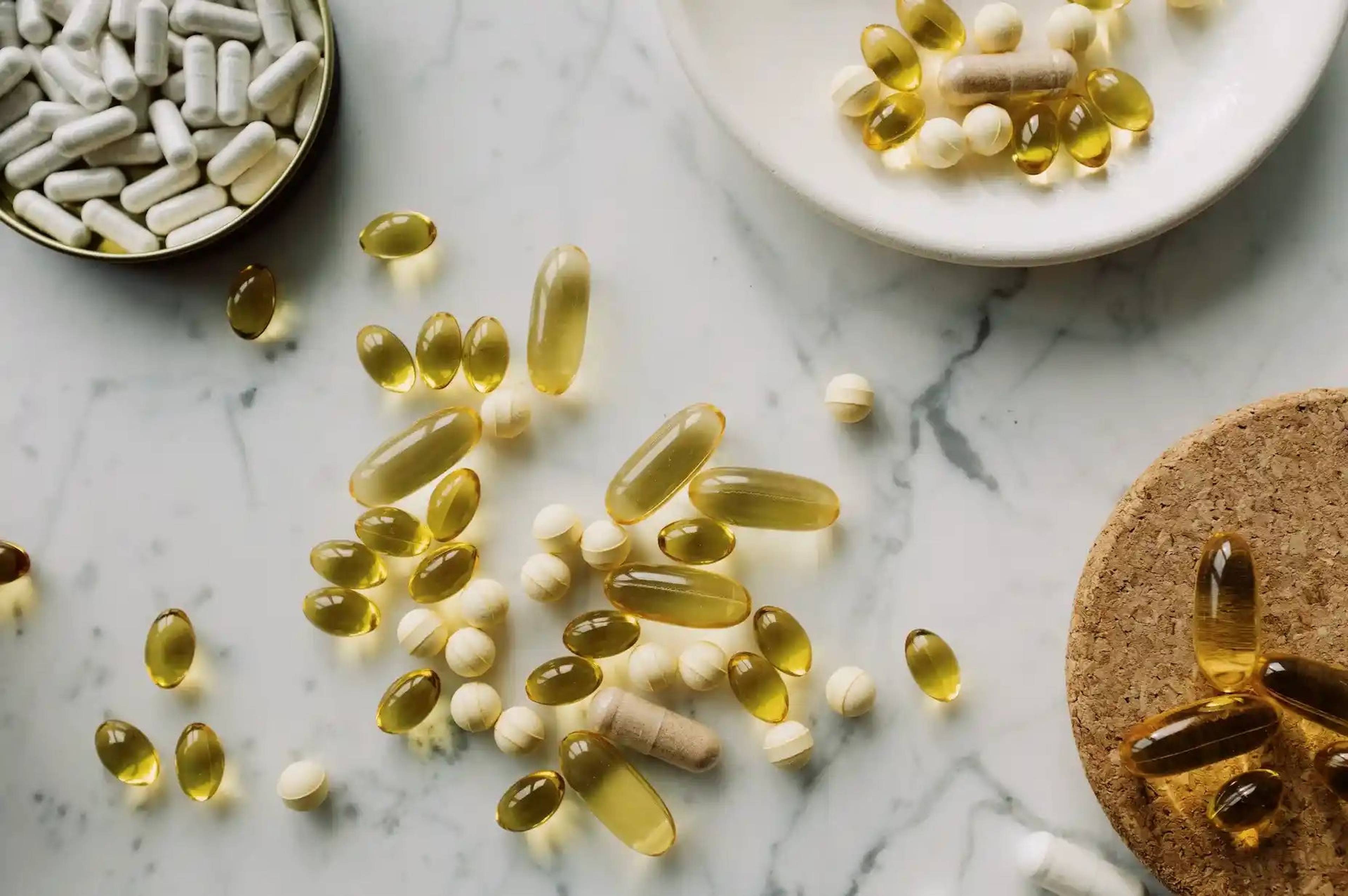Assorted capsules, tablets, and golden softgels scattered on a white marble surface beside small dishes.