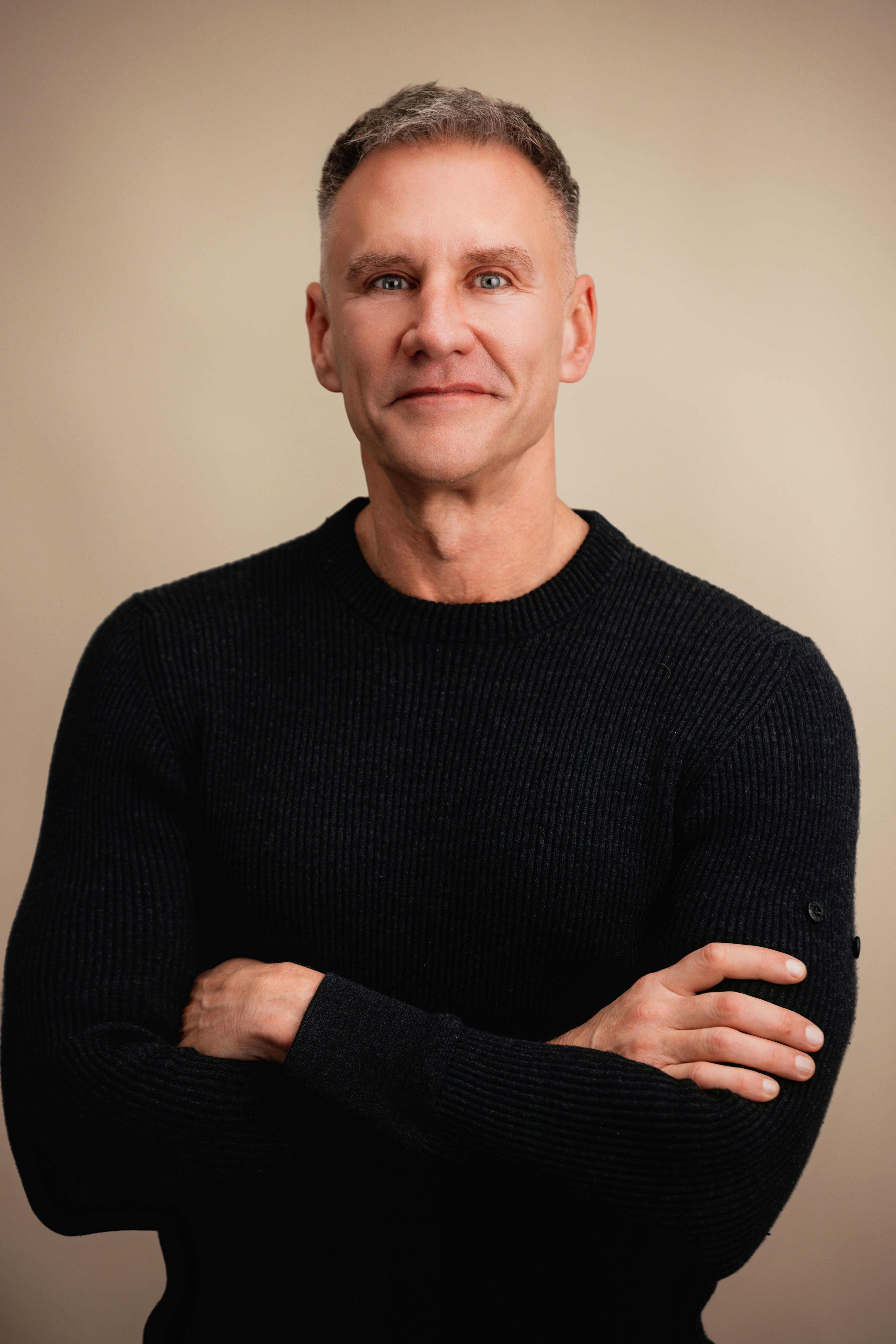 Portrait of a man in a black sweater with his arms crossed against a beige background.