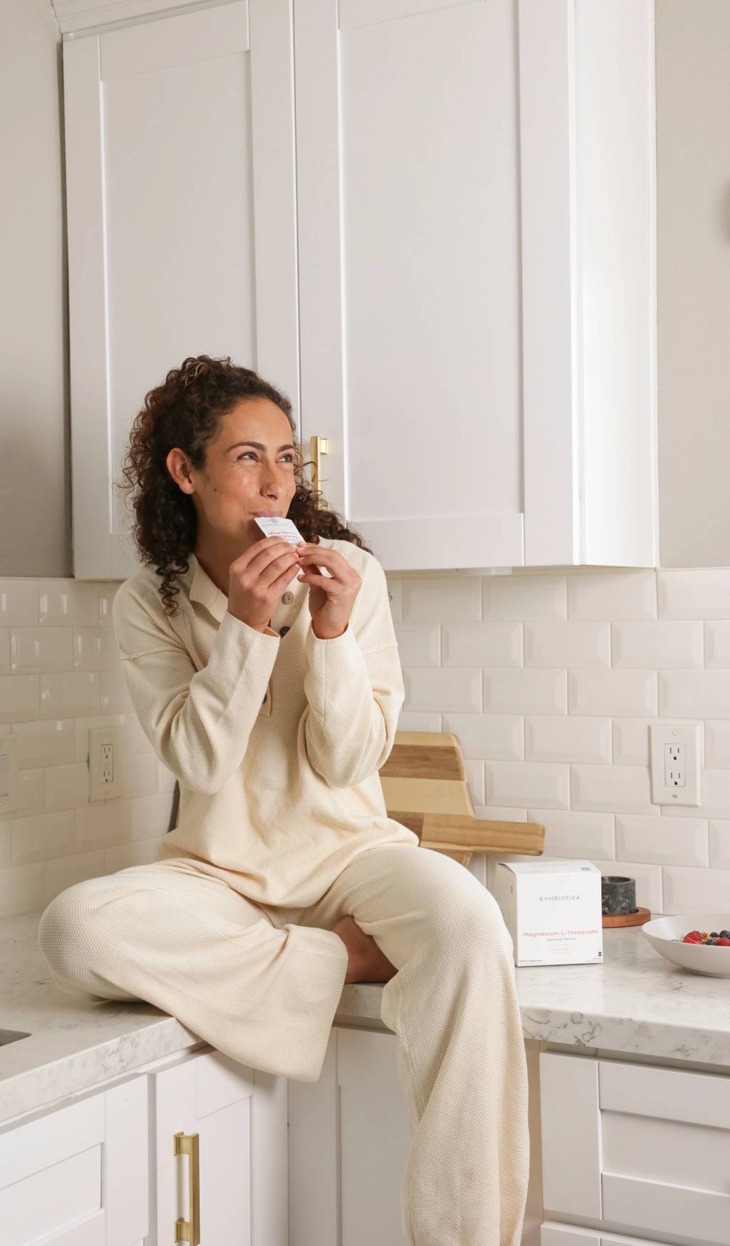 Woman in cream loungewear sits on a kitchen counter holding a small packet near her mouth, with a supplement box beside a bowl of berries.