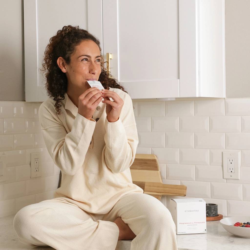 Woman in cream loungewear sits on a kitchen counter holding a small packet near her mouth, with a supplement box beside a bowl of berries.