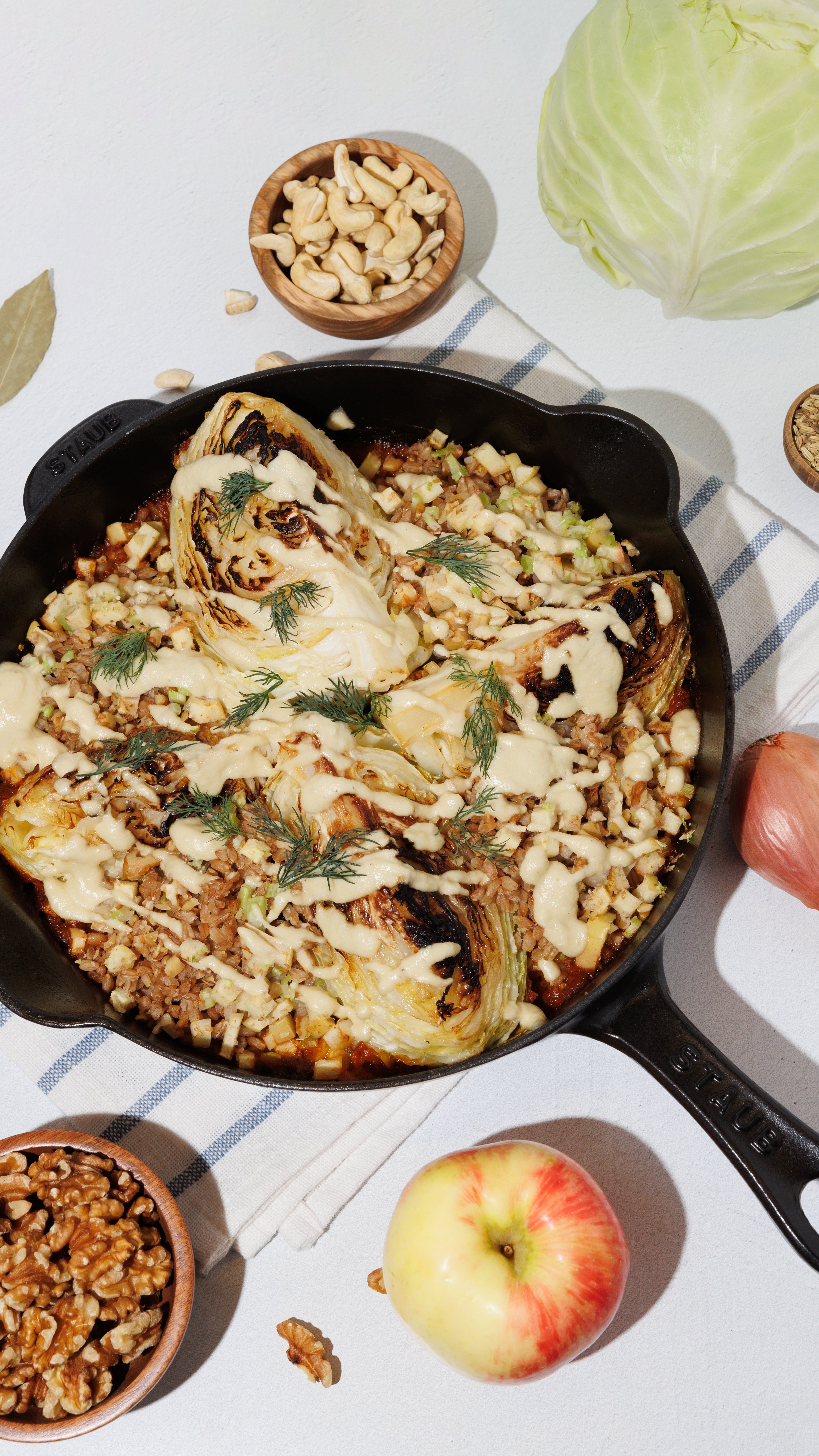Cabbage wedges with creamy sauce and herbs in a skillet, with bowls of cashews and walnuts and an apple nearby