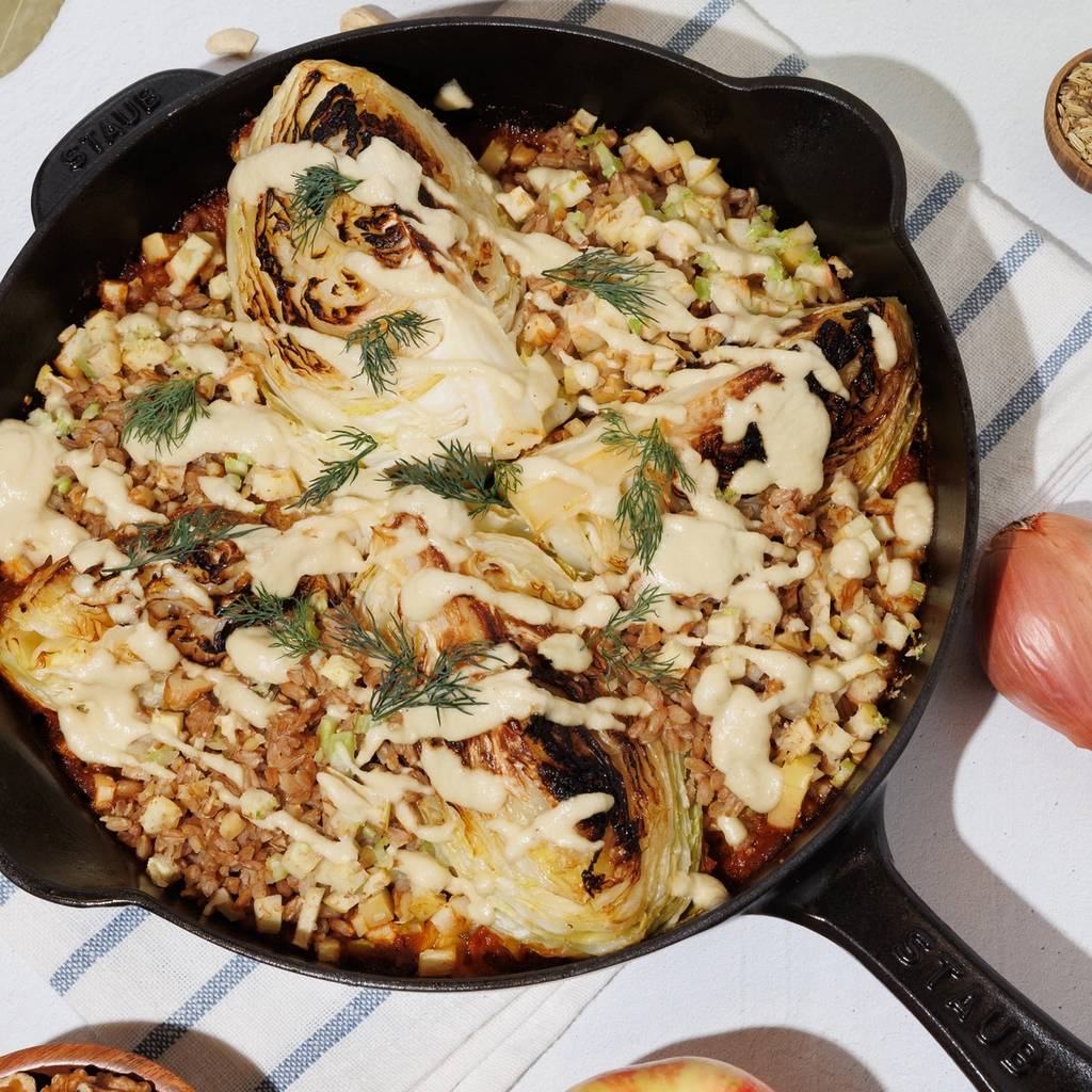 Cabbage wedges with creamy sauce and herbs in a skillet, with bowls of cashews and walnuts and an apple nearby
