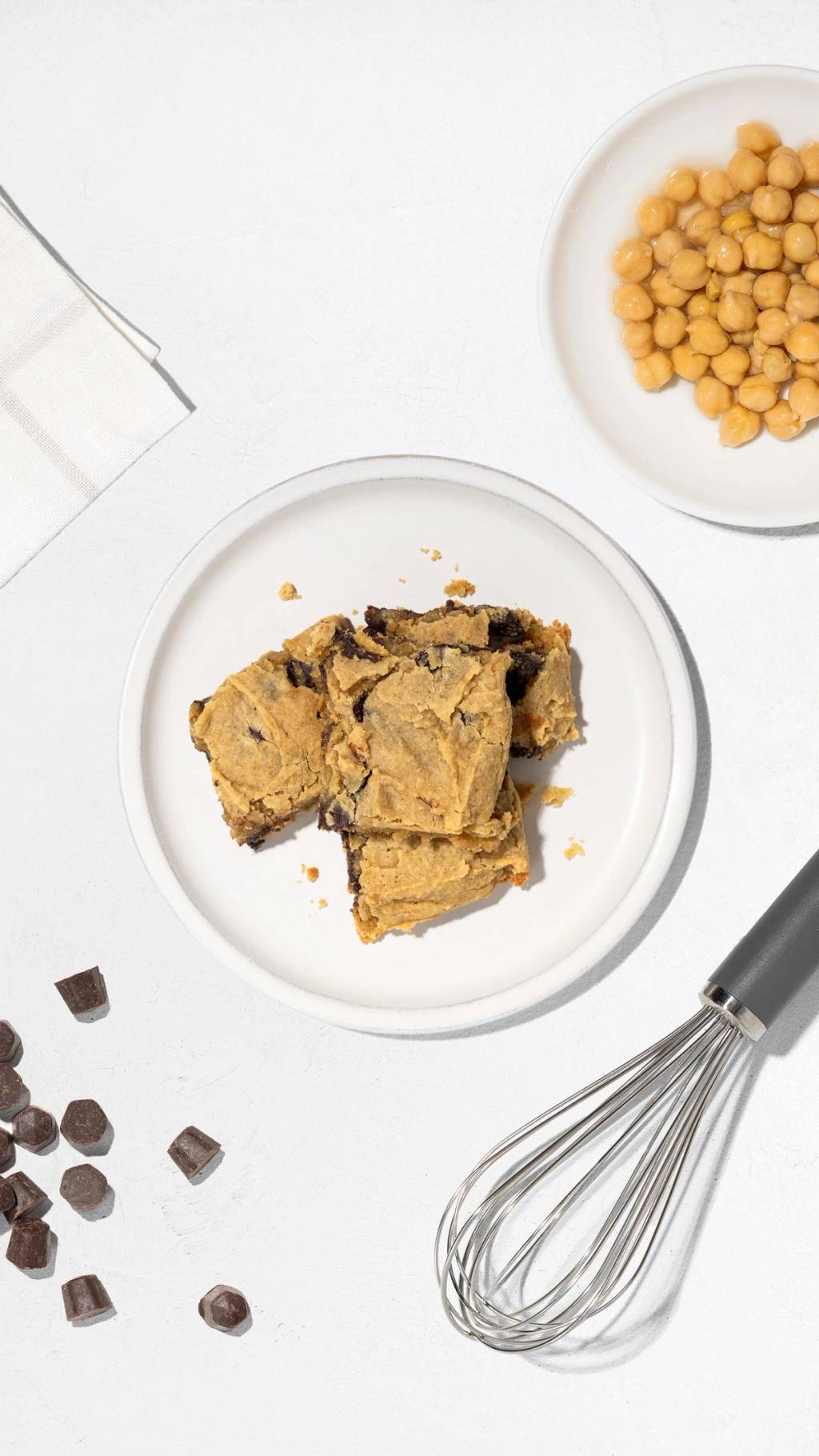 Plate of chickpea blondies with chickpeas, chocolate chunks, and a whisk on a white surface.