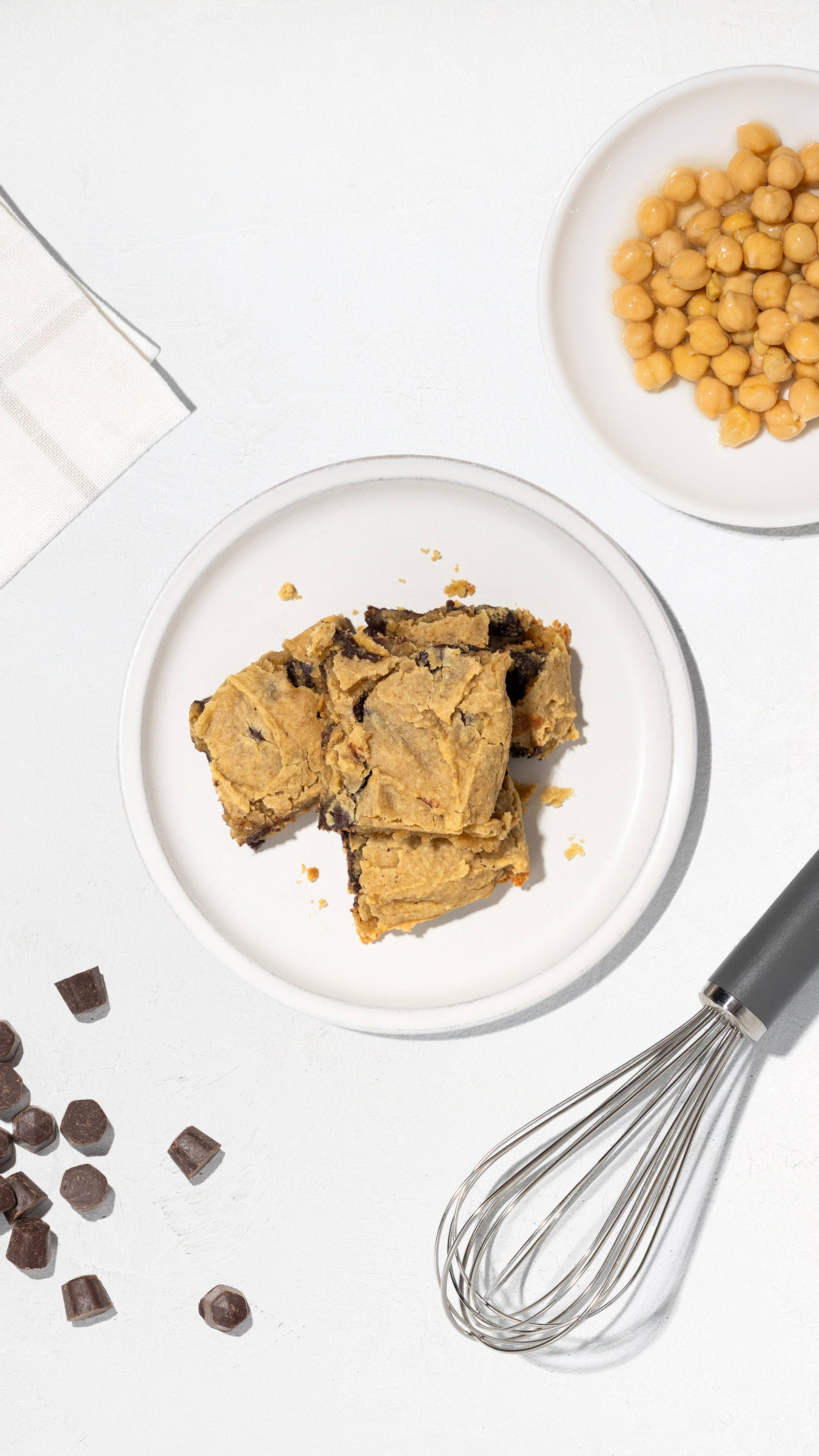 Plate of chickpea blondies with chickpeas, chocolate chunks, and a whisk on a white surface.