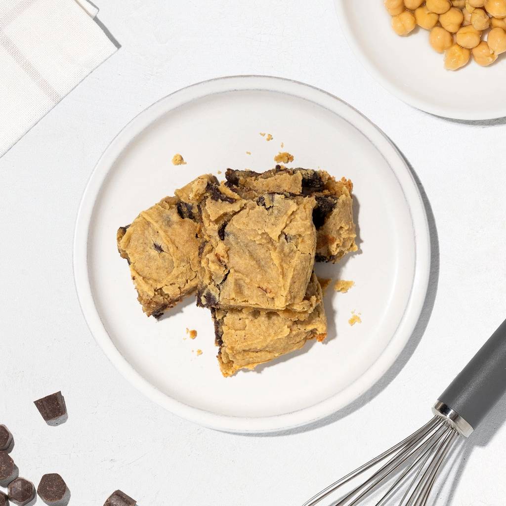 Plate of chickpea blondies with chickpeas, chocolate chunks, and a whisk on a white surface.