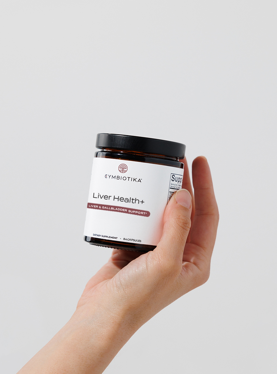 A hand holds a jar labeled Cymbiotika Liver Health+.