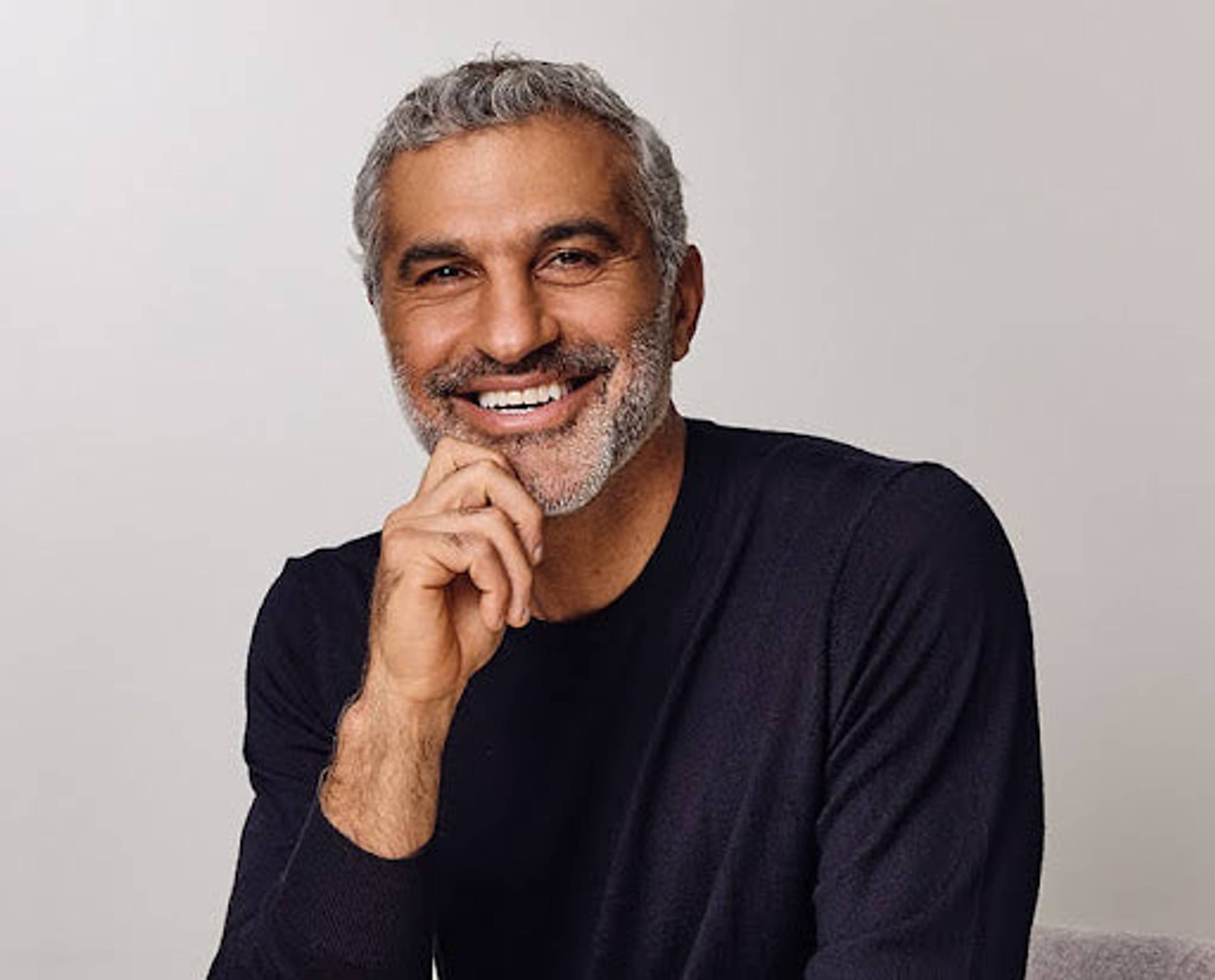 Smiling gray-haired man in a dark sweater resting his hand beneath his chin.