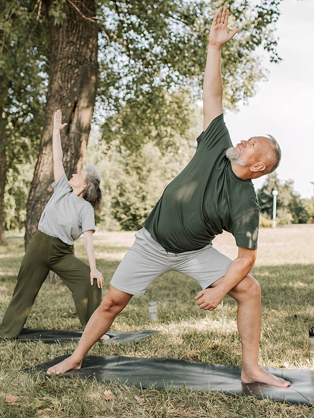 Older man and woman doing side stretches on yoga mats in a park.
