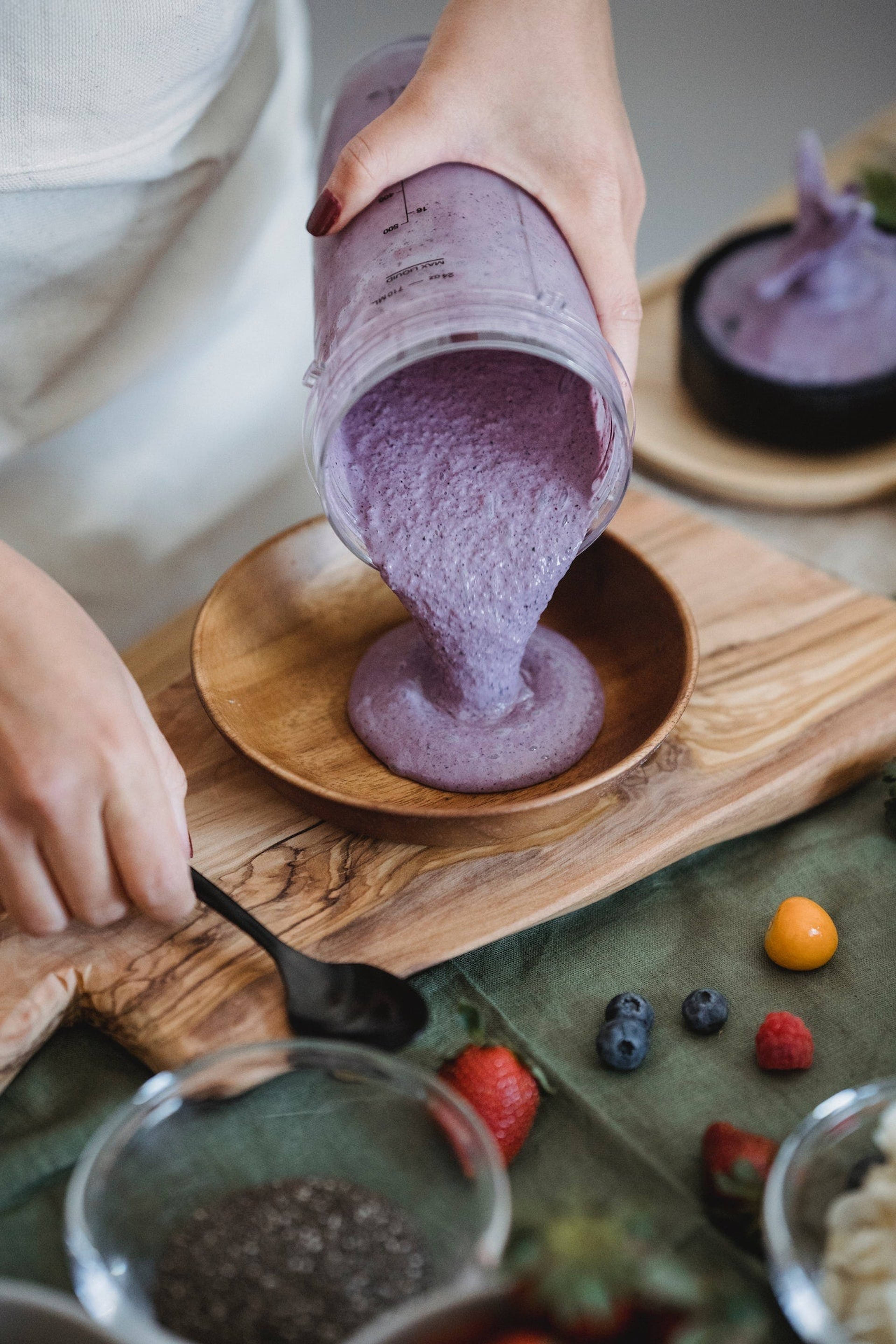Hands pour a thick purple smoothie from a marked blender cup into a wooden bowl, with berries and small bowls nearby.