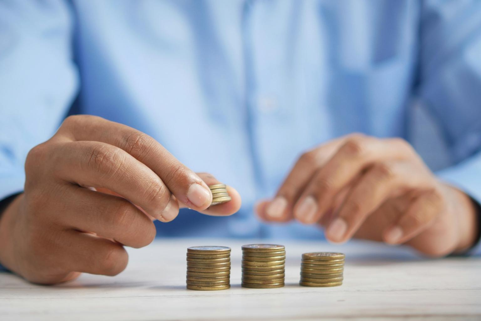 A person in a blue shirt stacks coins into increasing piles, symbolizing careful financial management and incremental investment growth.