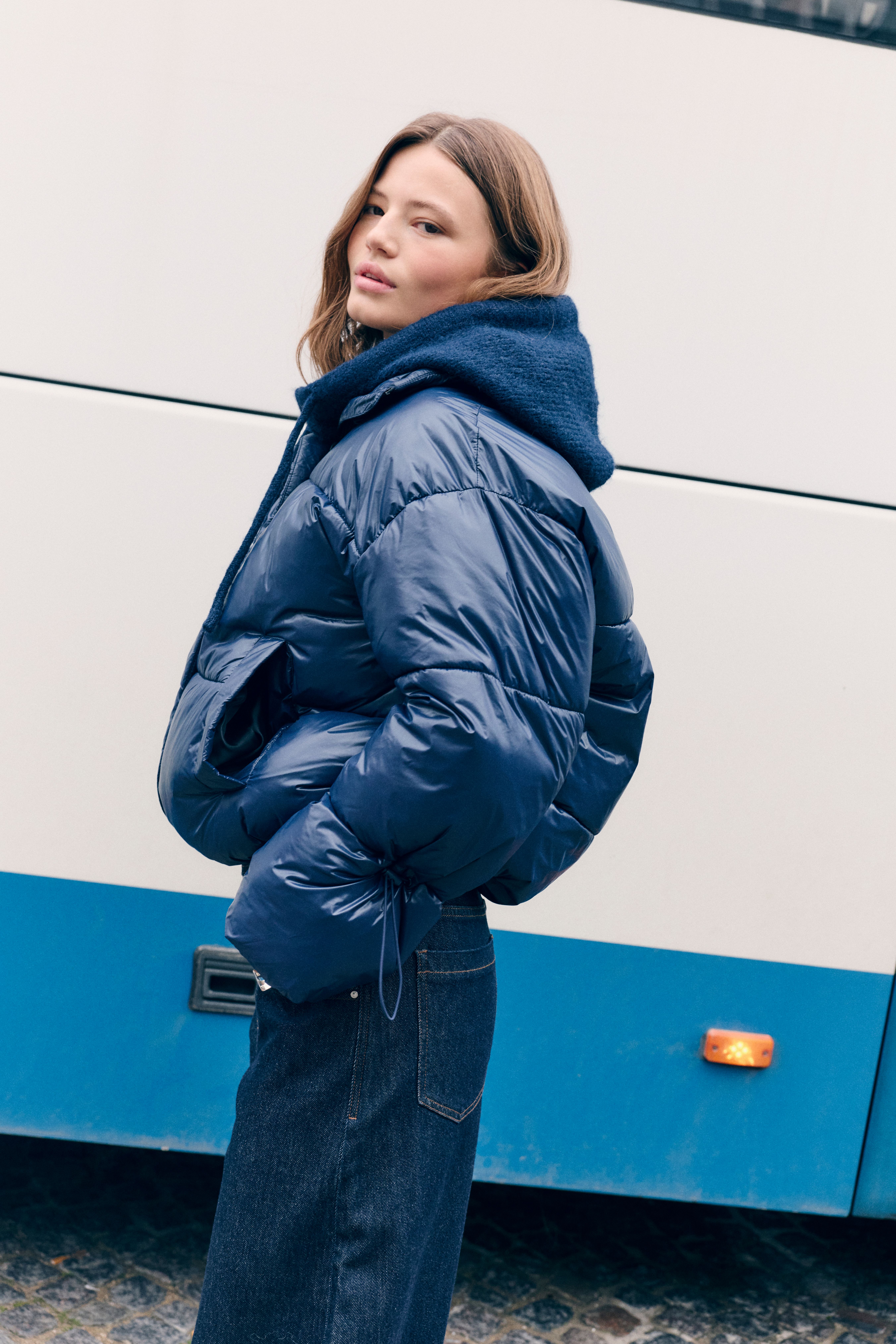 Donna con piumino blu e jeans scuri guarda indietro su una strada urbana accanto a un bus