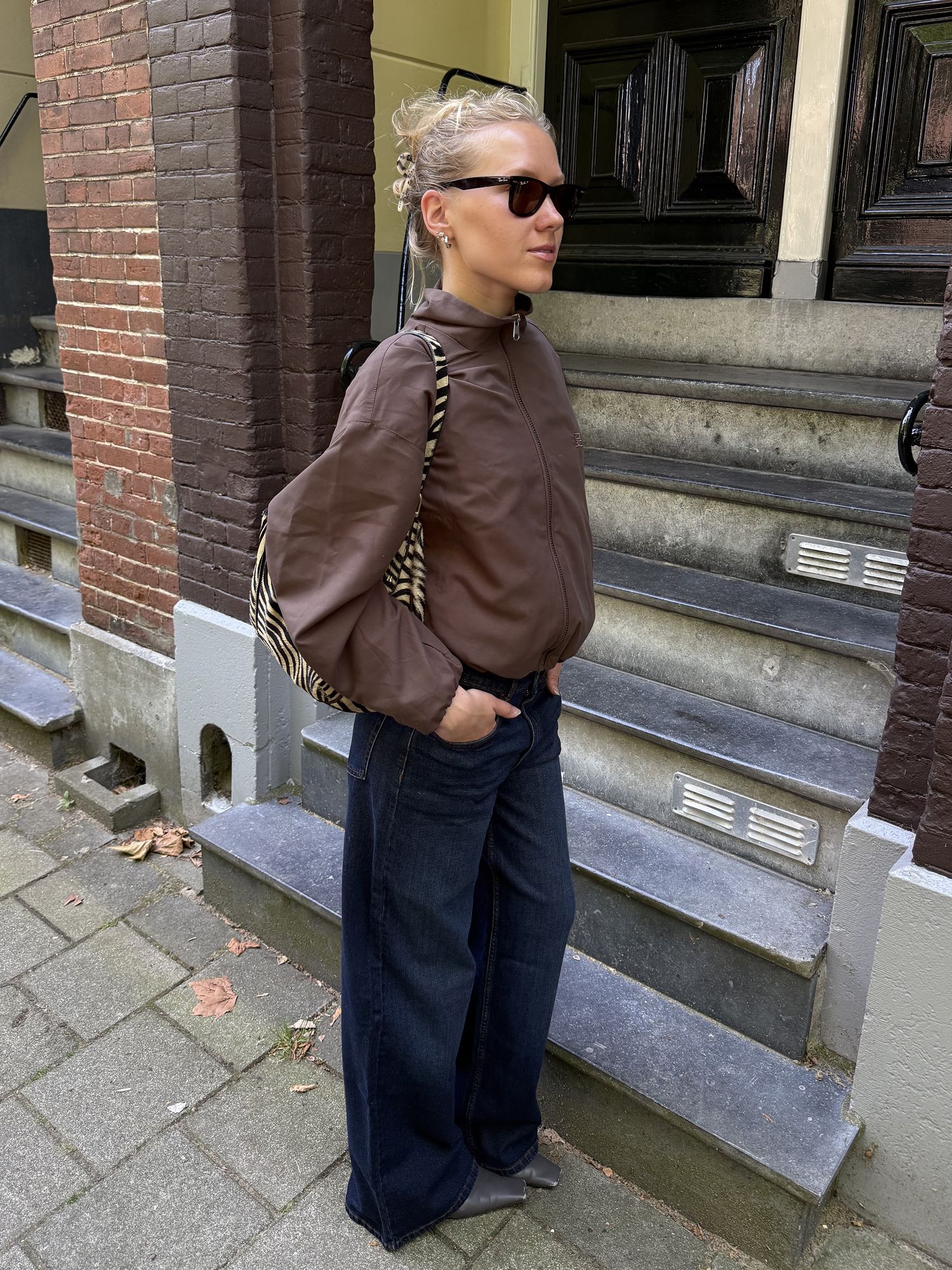 “Woman wearing a brown zip-up jacket, dark wide-leg jeans and sunglasses.