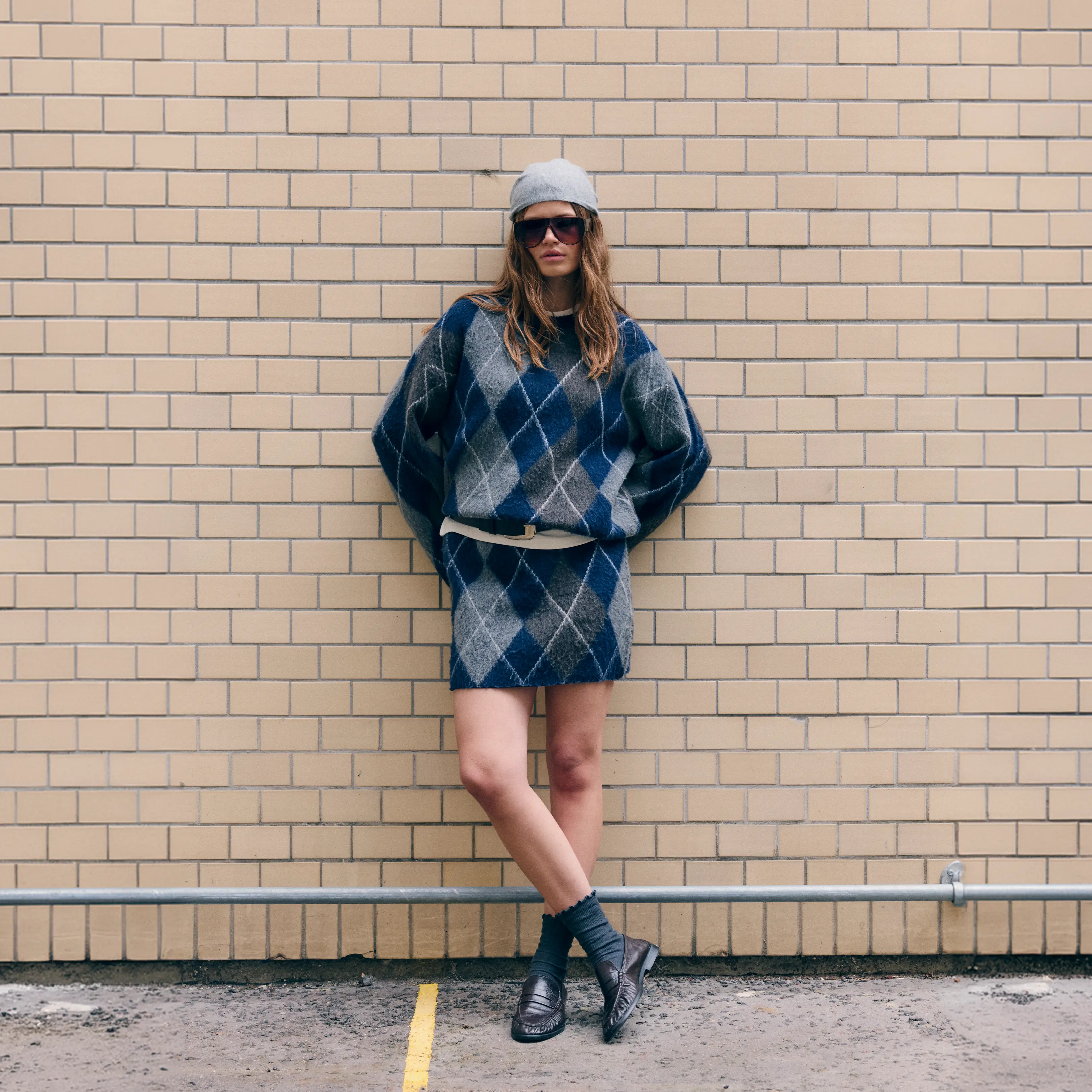 Woman in blue-grey argyle set and beanie leans against a brick wall, legs crossed.
