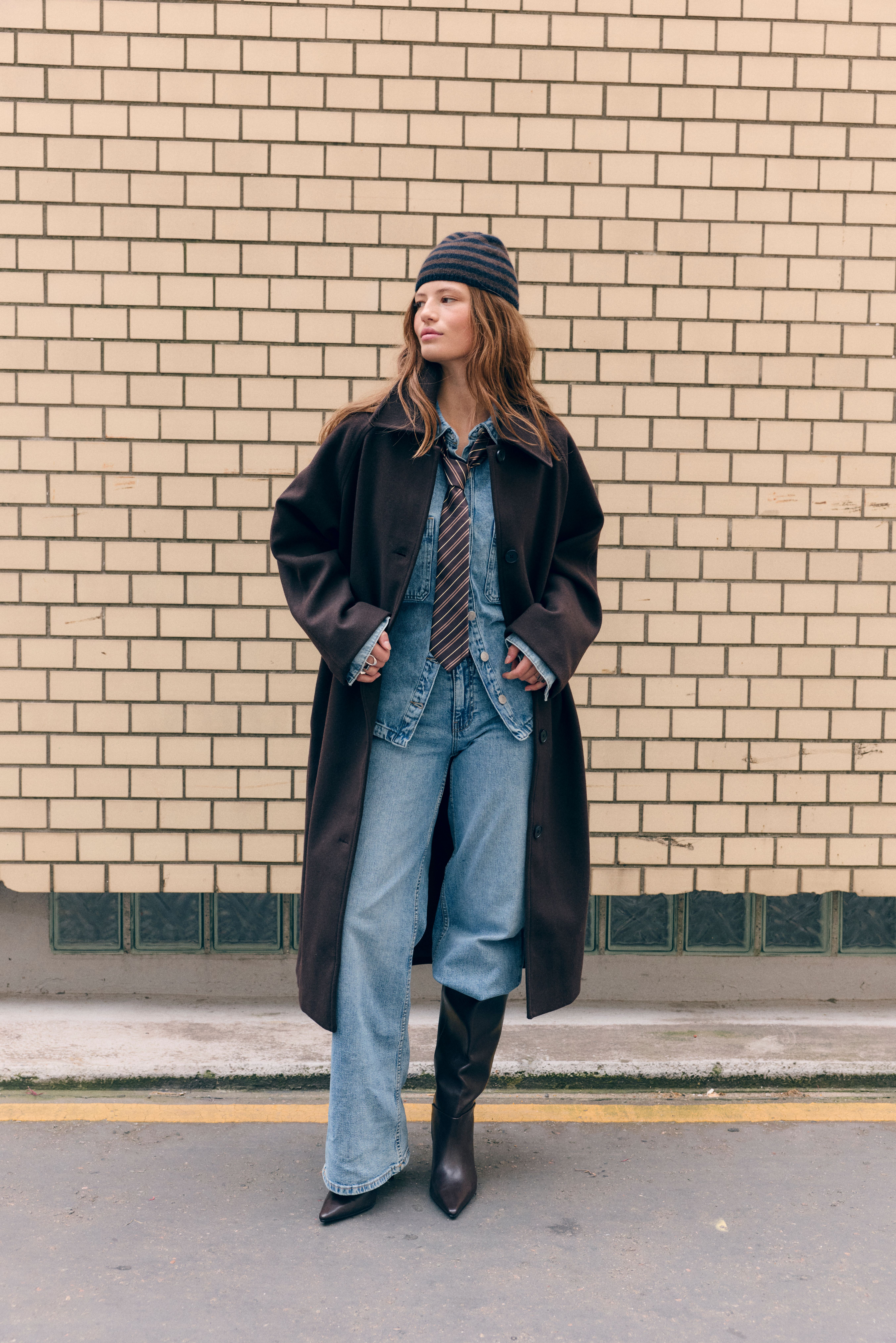 Donna in cappotto marrone e jeans larghi davanti a un muro di mattoni