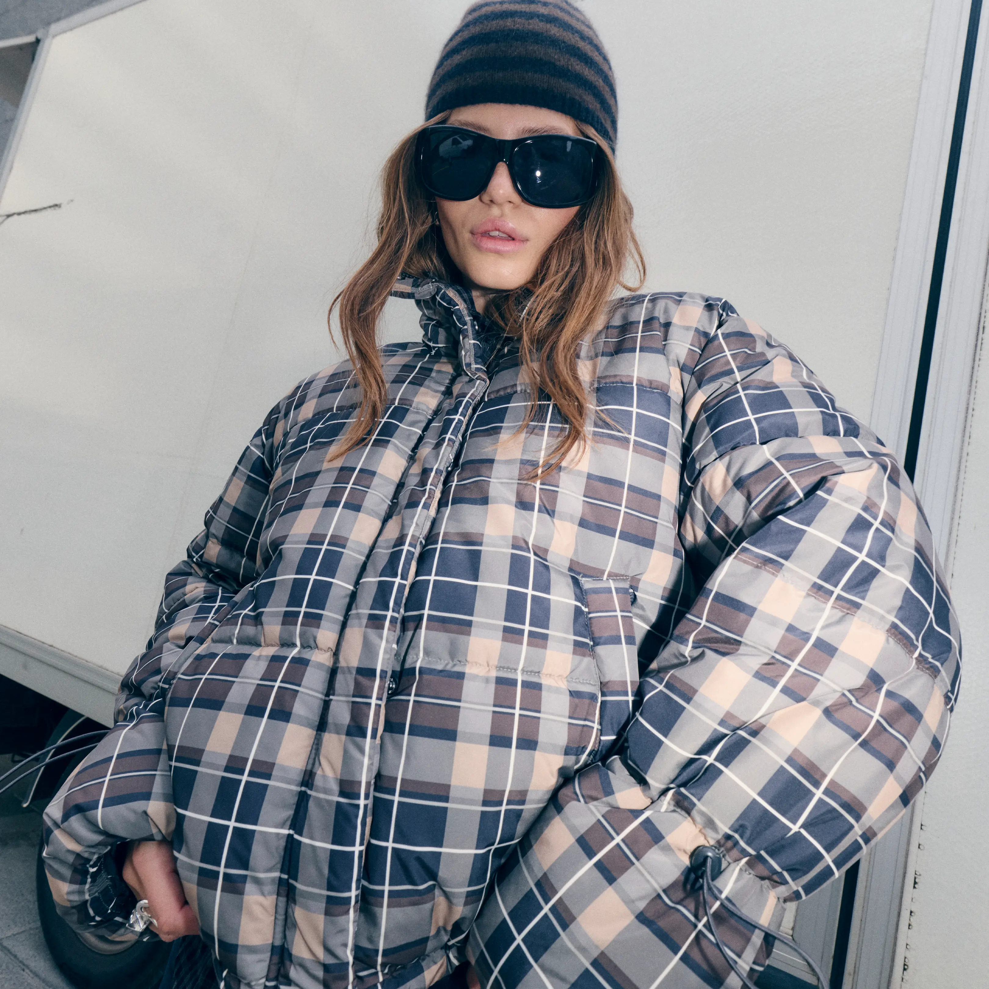 Woman in plaid puffer jacket, striped beanie, and oversized sunglasses looking at camera
