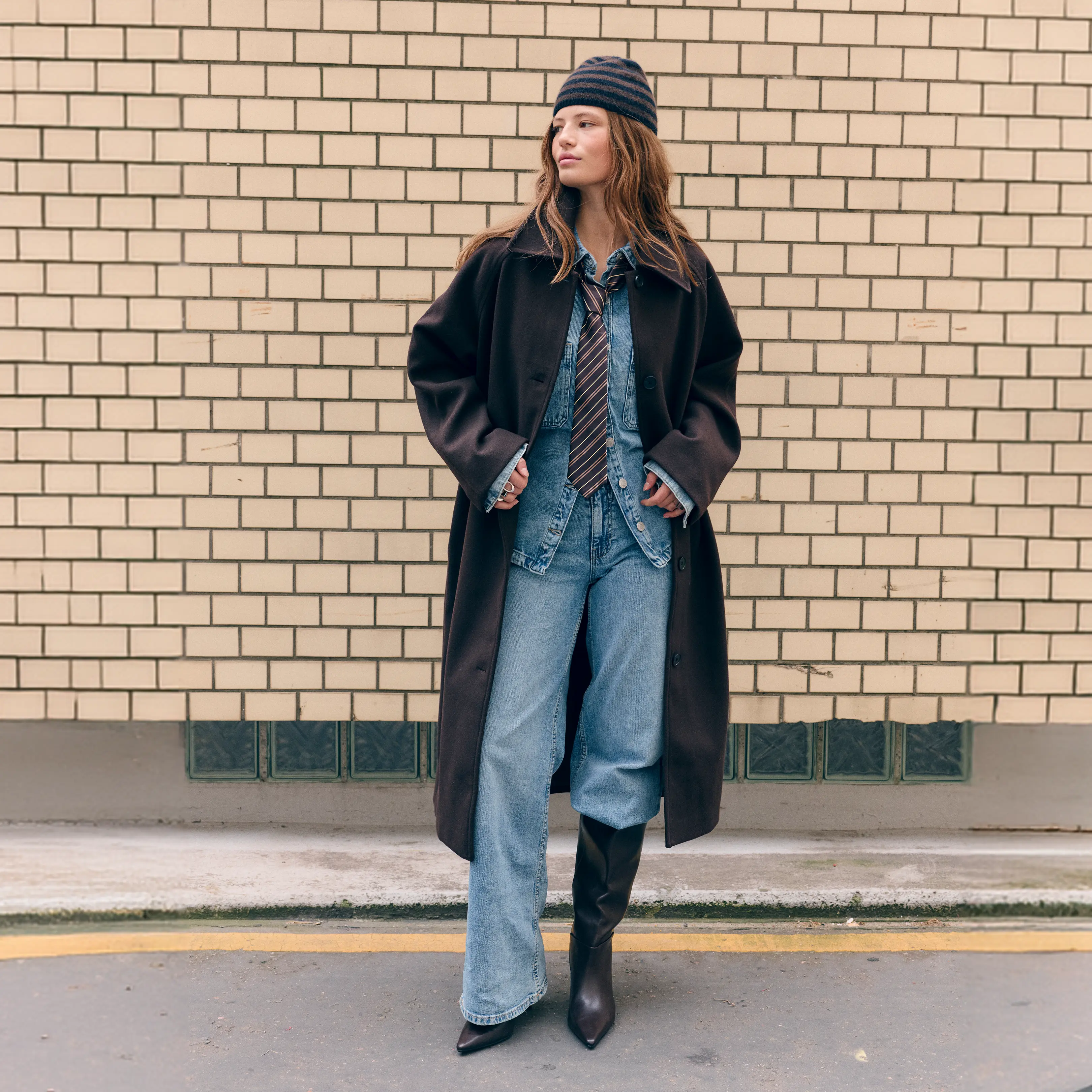 Woman in brown coat, denim outfit, striped beanie and tie, standing by a brick wall.