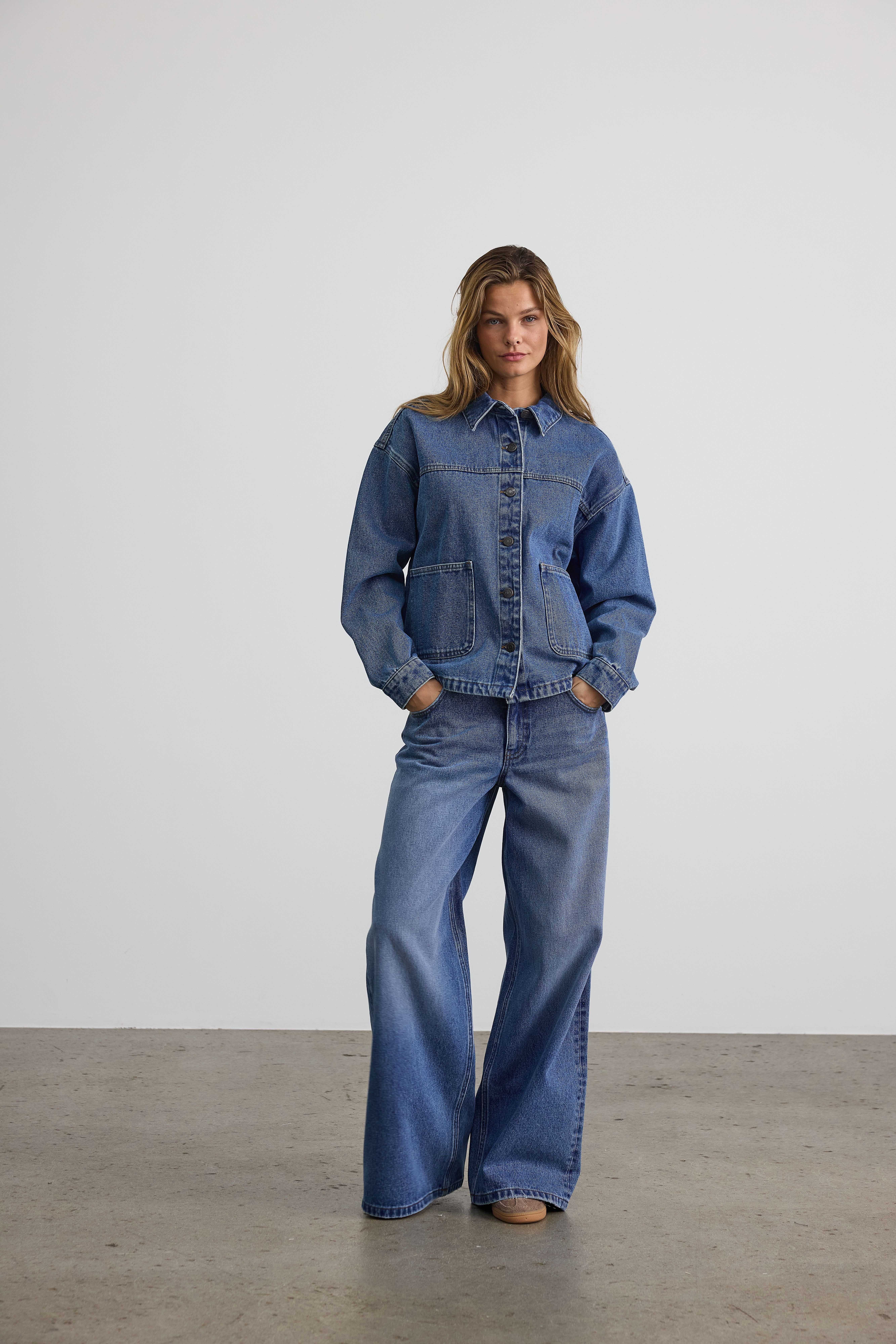 Vrouw in oversized spijkerjas en wijde jeans, staand met handen in zakken op wit.