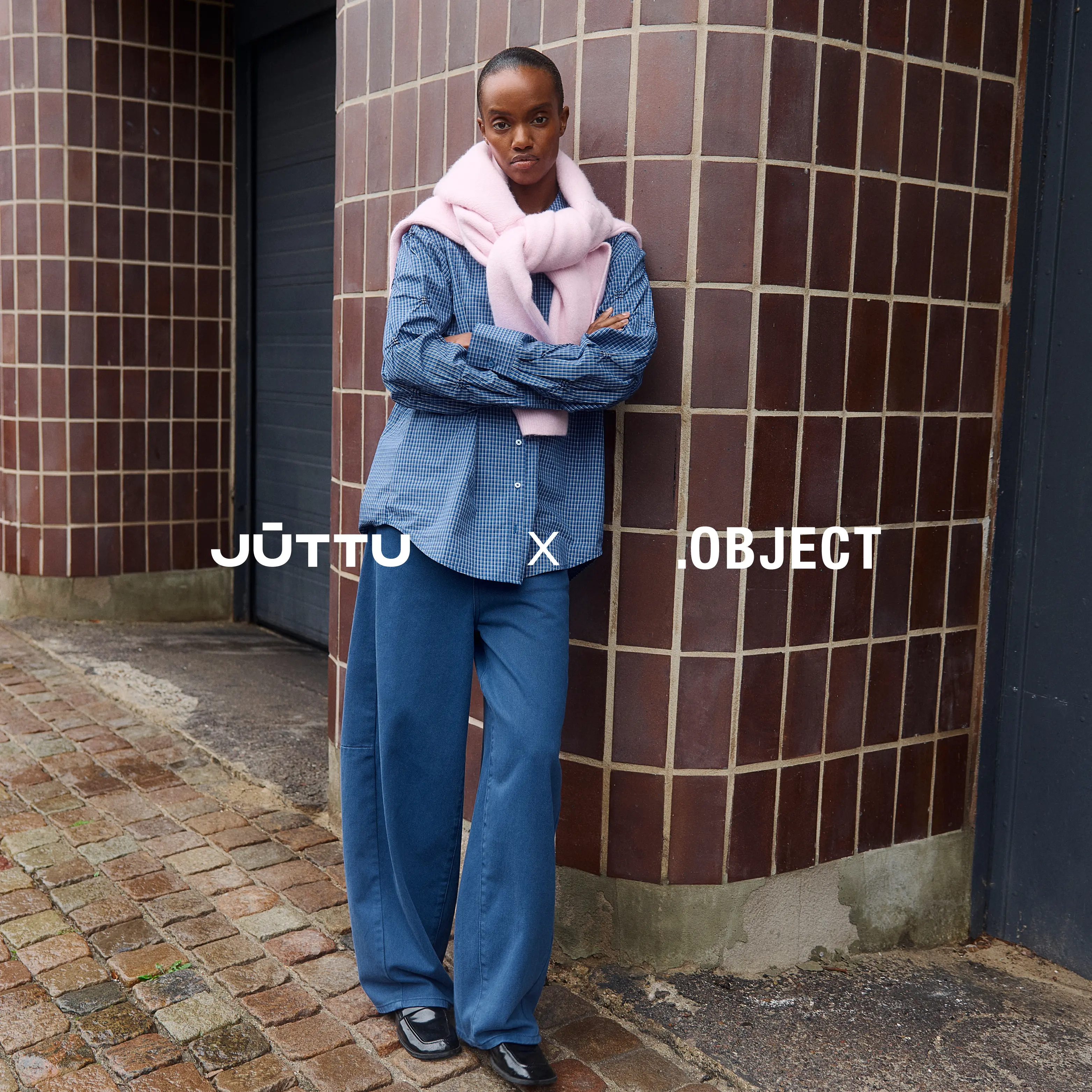 Kvinde i blåternet skjorte, jeans og pink tørklæde læner sig mod flisevæg med korslagte arme