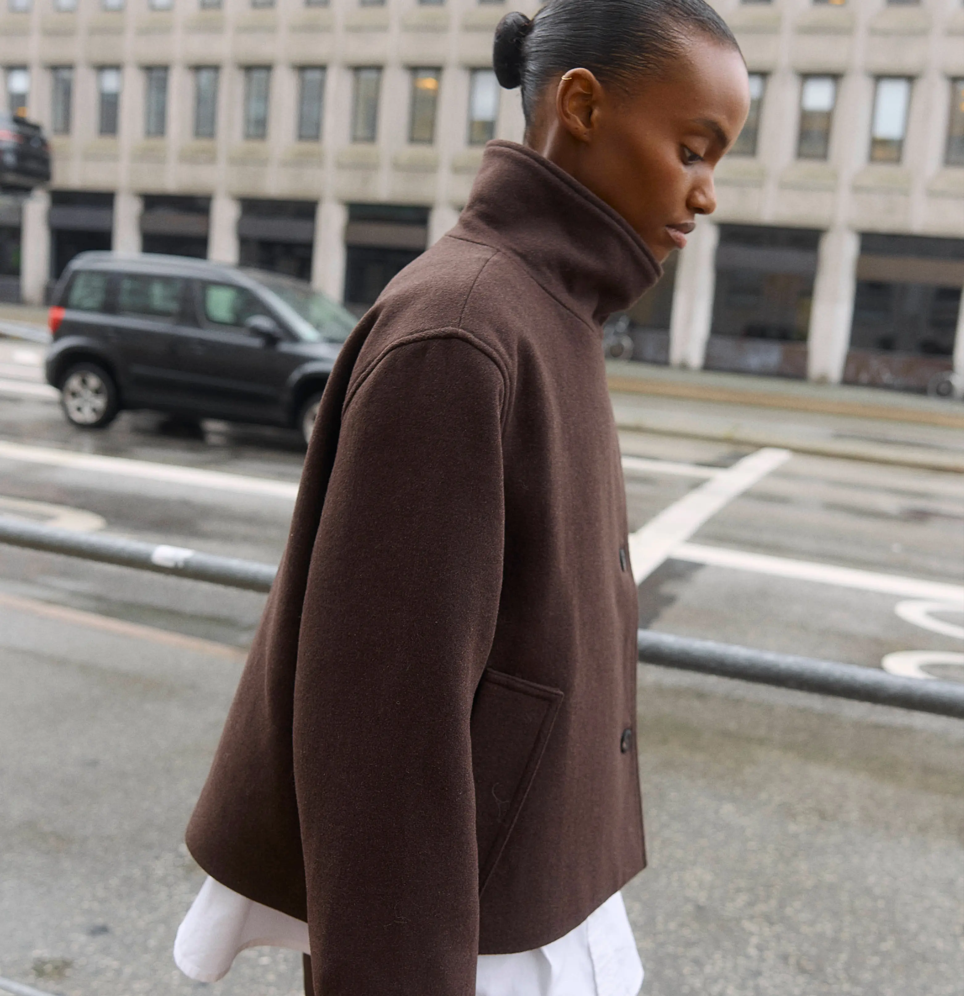 Woman in a brown cropped jacket walking along a city street in profile.