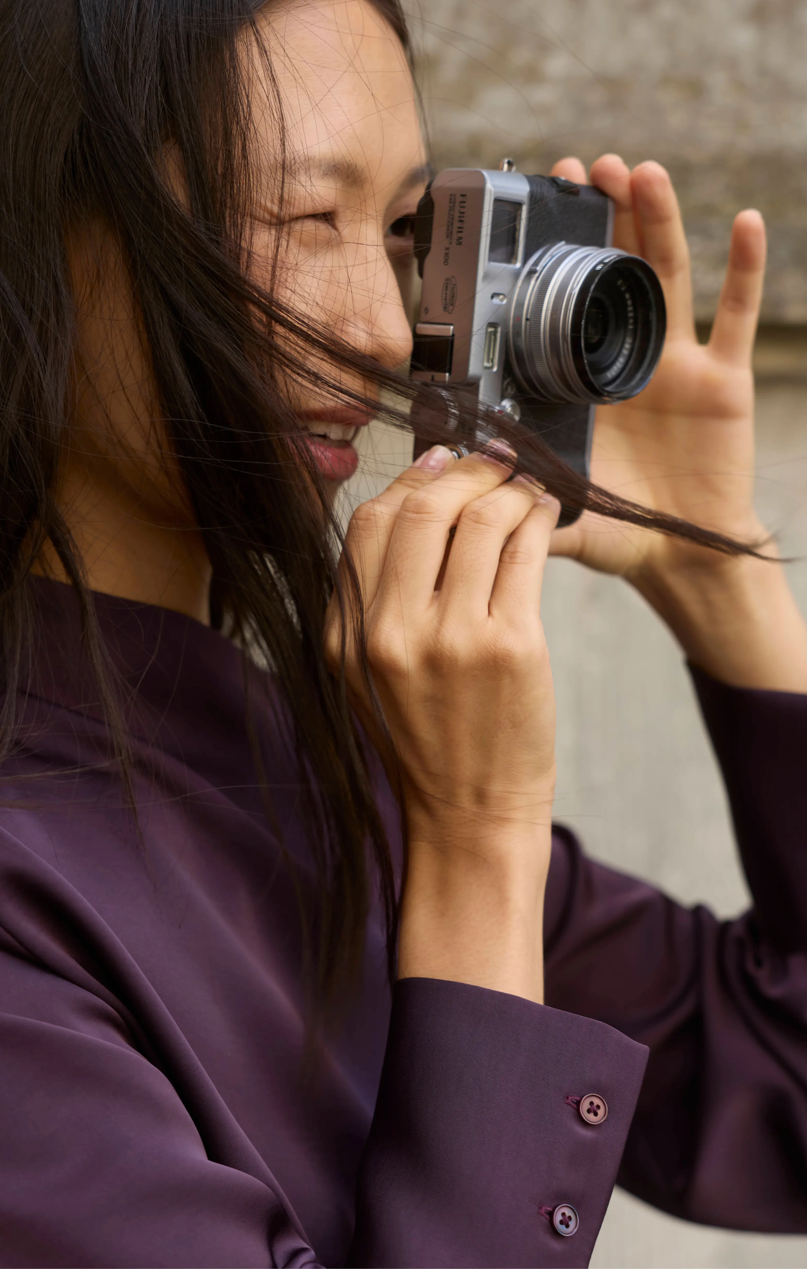 Smiling woman in a purple shirt taking a photo with a Fujifilm mirrorless camera outdoors