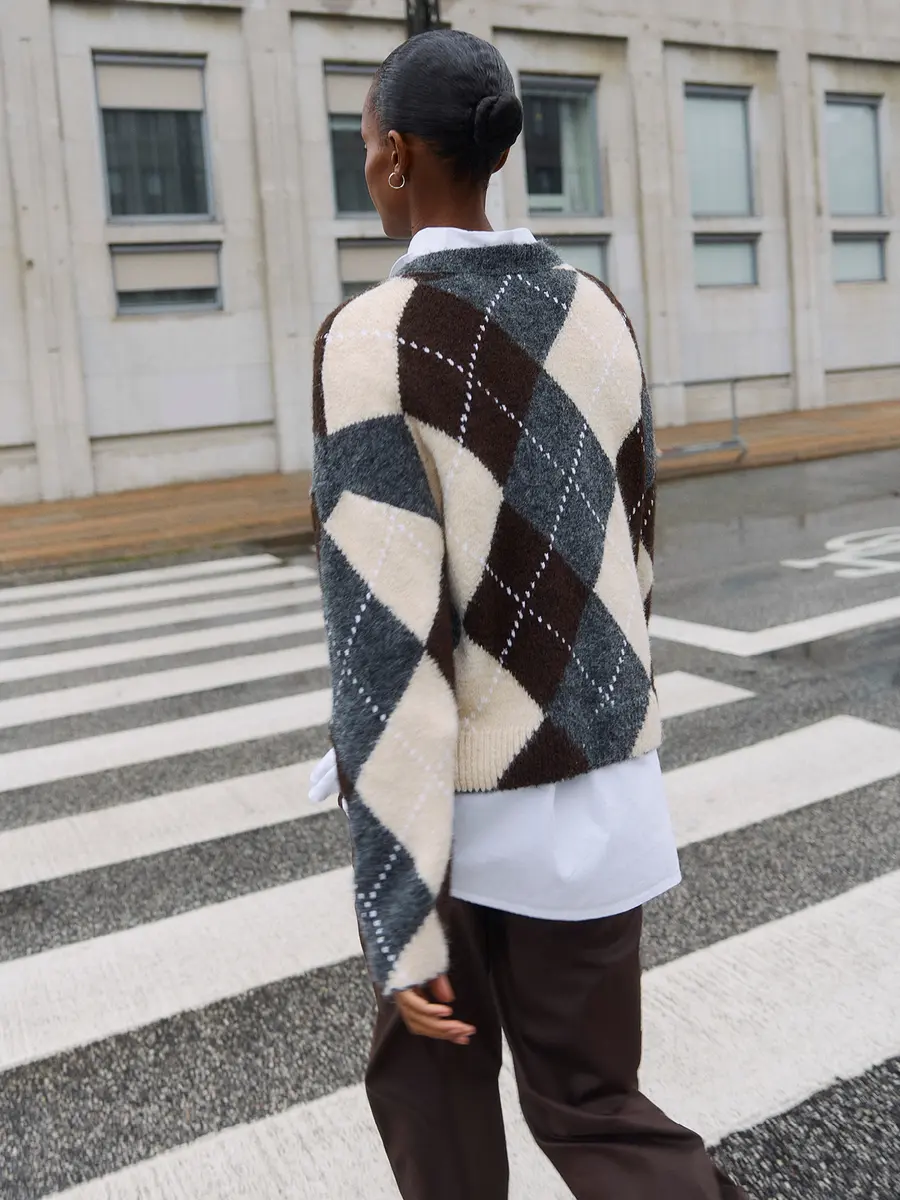 Woman in an argyle sweater and brown pants walks across an urban crosswalk, seen from behind.