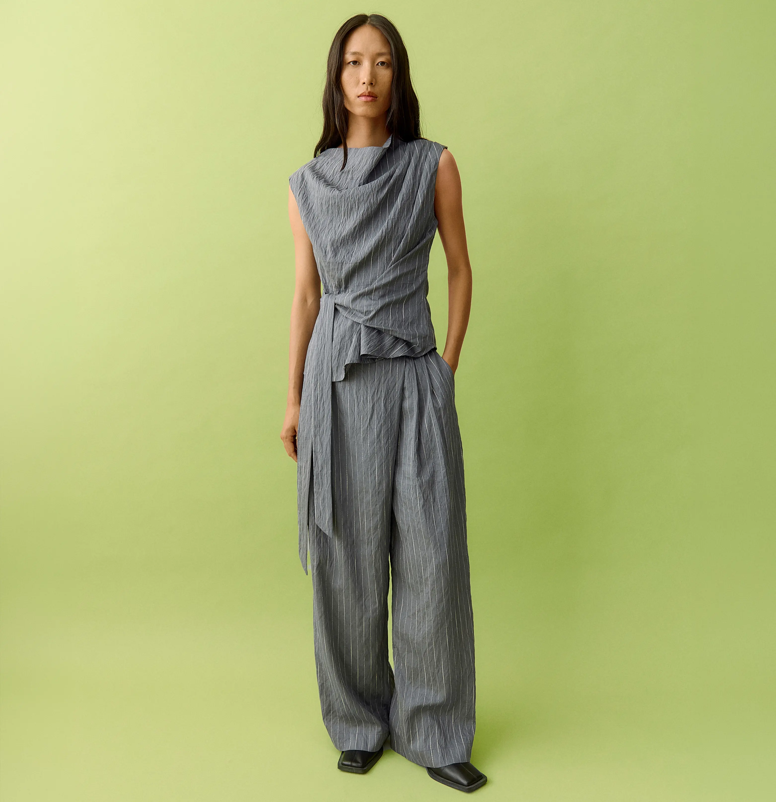 Woman in draped top and gray pinstriped wide-leg pants on green background