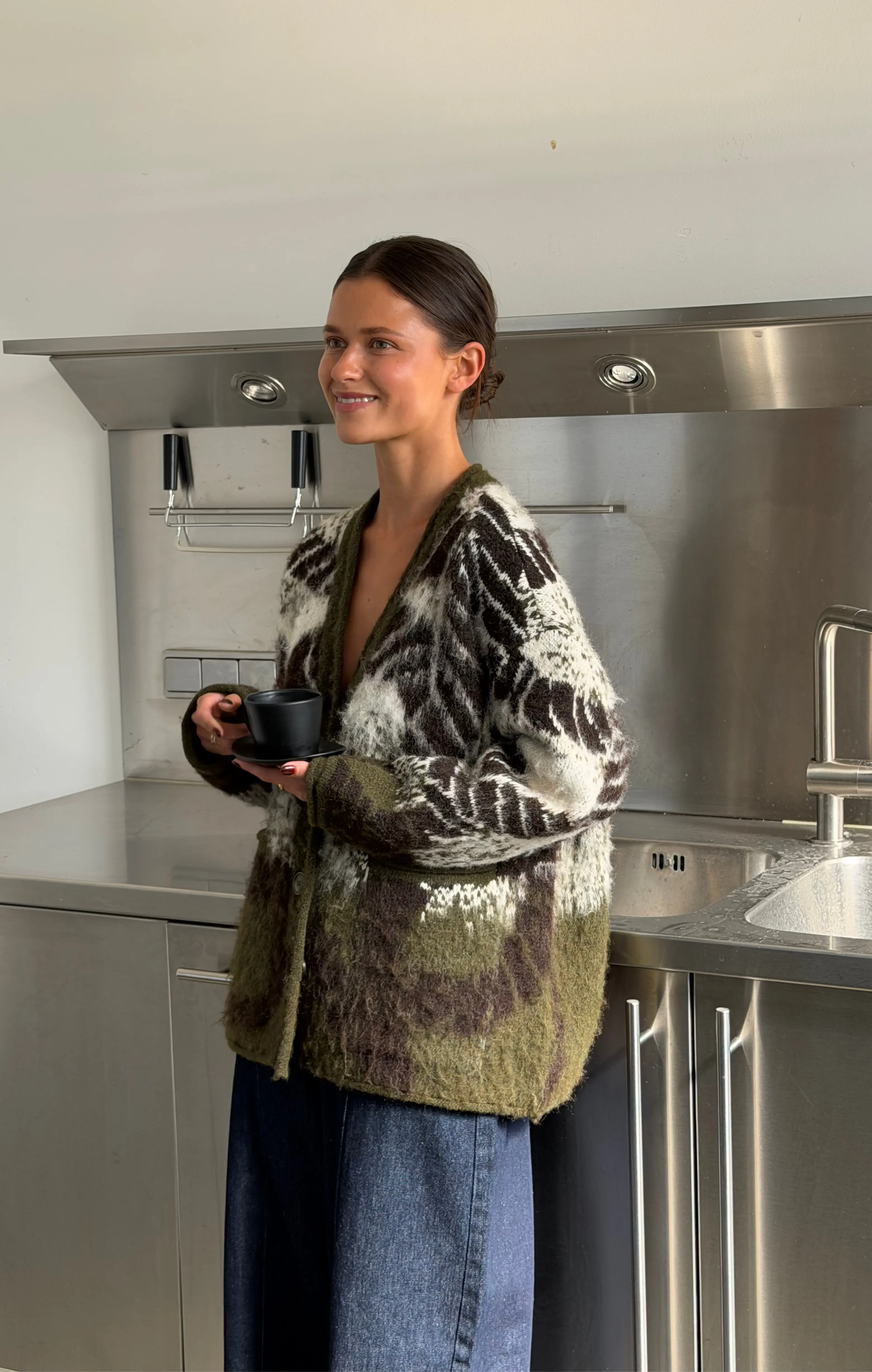 Vrouw met groene gebreide cardigan en kopje in minimalistische roestvrijstalen keuken