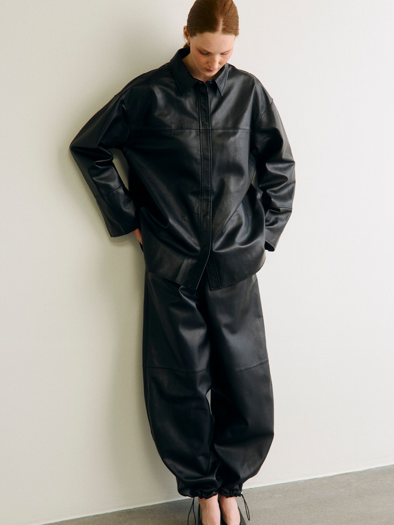 Kvinnlig modell i oversize svart läderoutfit lutar mot en vit vägg.