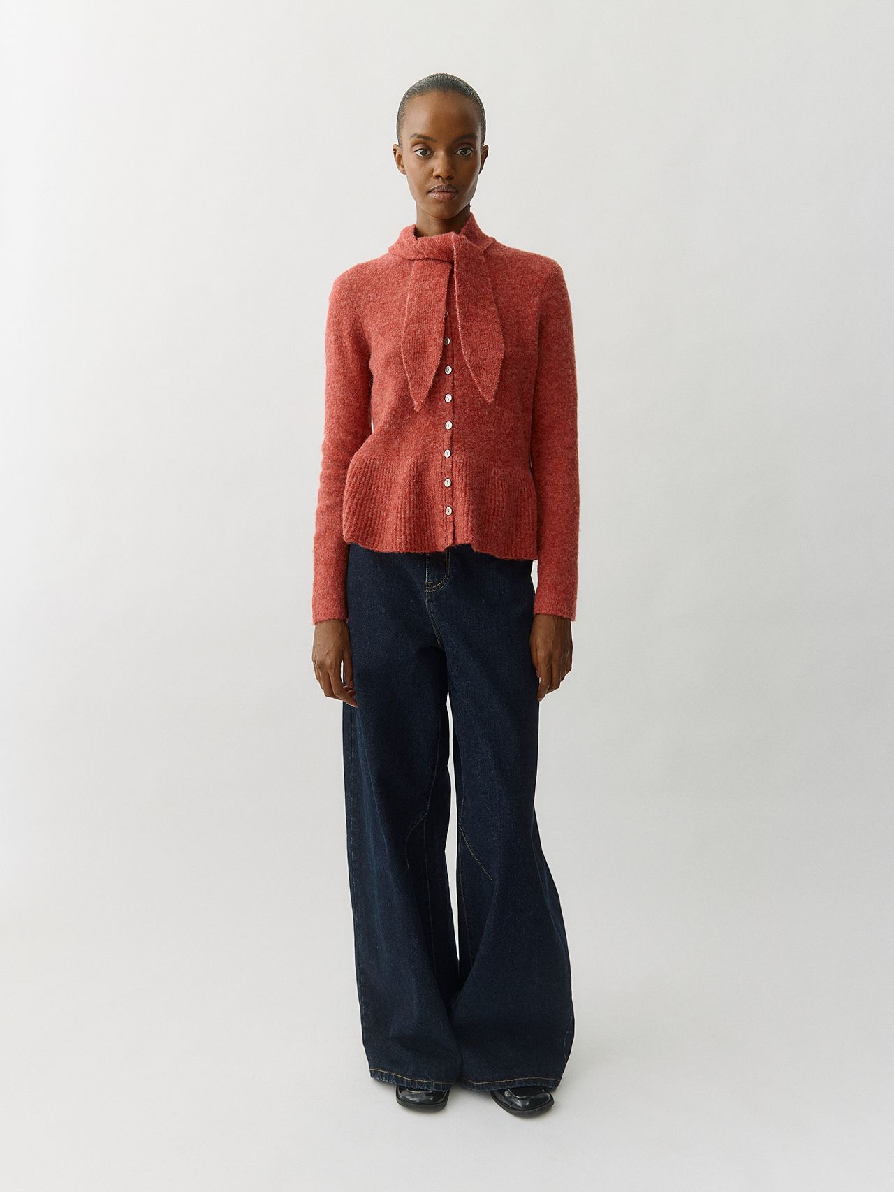 Woman in coral tie-neck cardigan and wide-leg dark jeans in a studio.