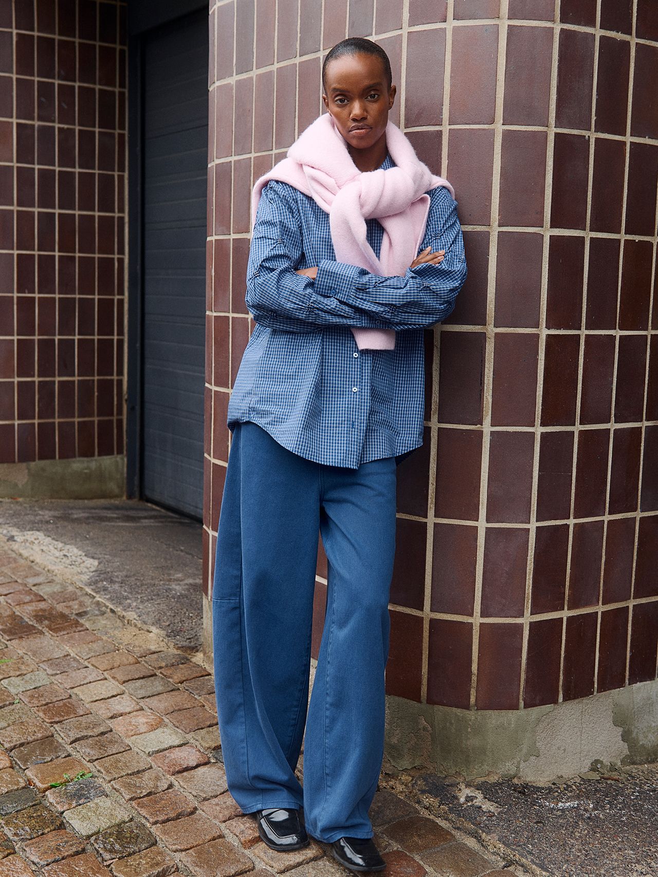 Woman in blue gingham shirt, pink sweater and wide-leg jeans by a tiled wall