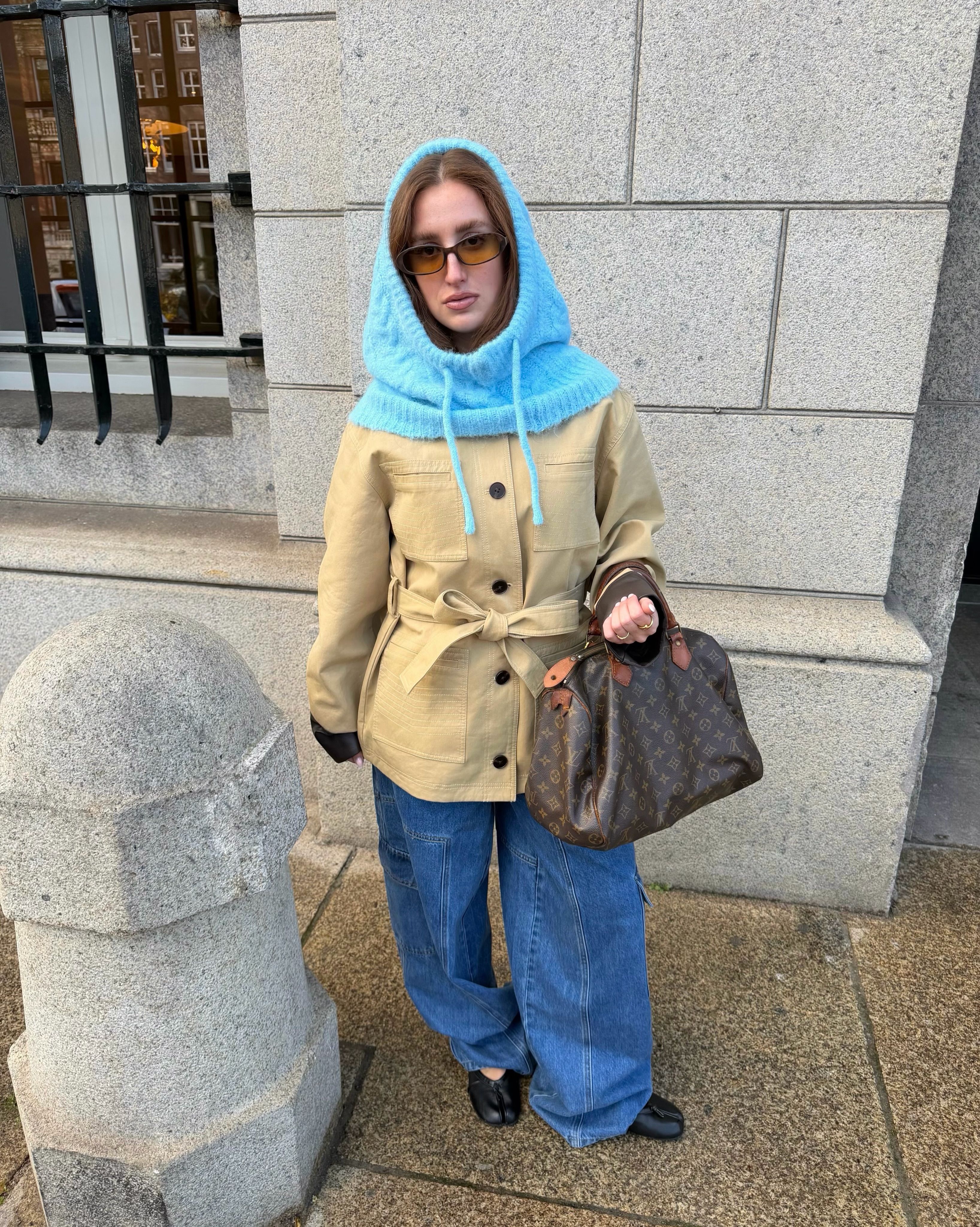 Frau mit hellblauer Strickhaube, beiger Jacke, weiten Jeans und Louis-Vuitton-Tasche.