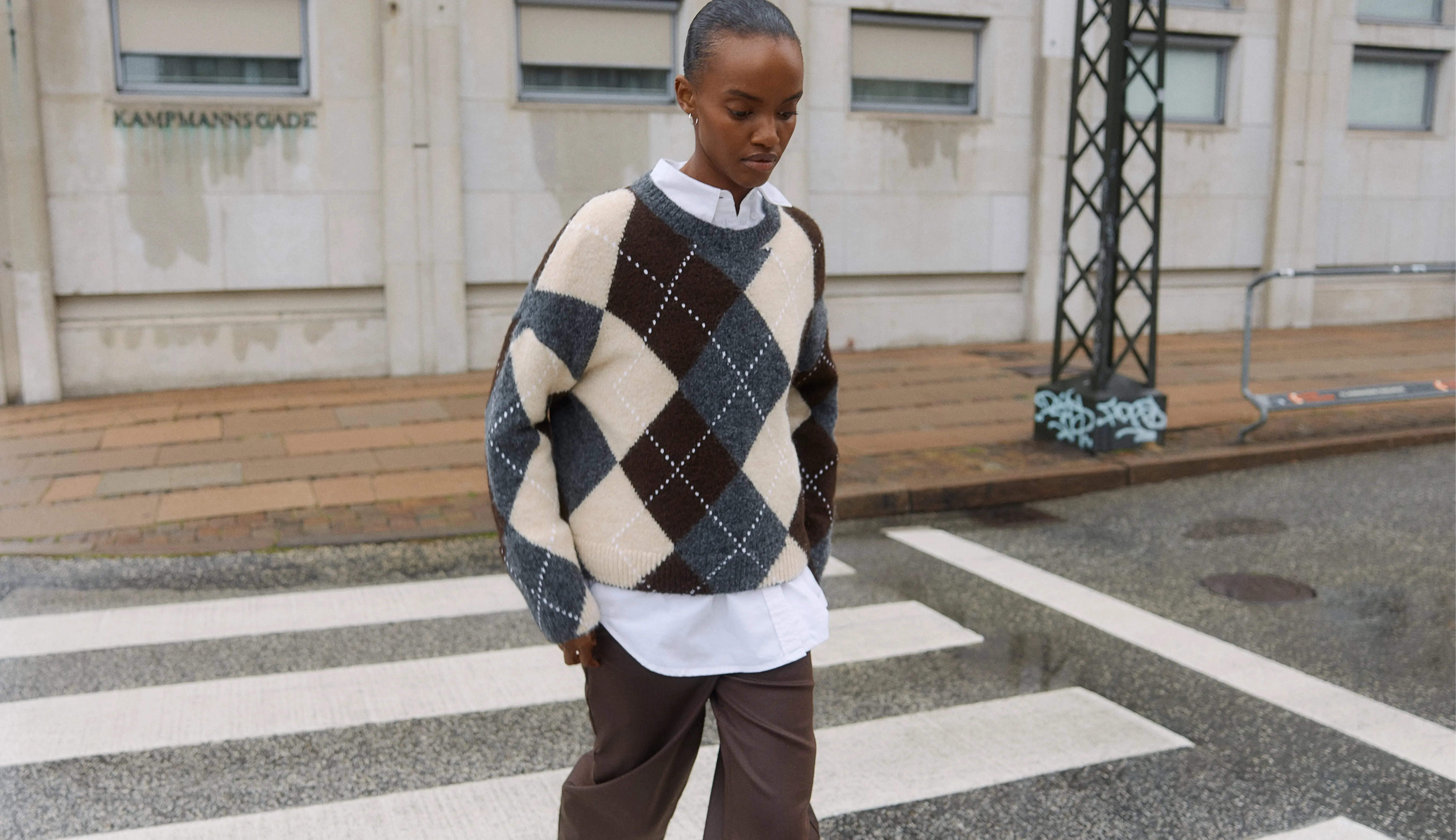 Woman in an argyle sweater and brown pants walking on a city crosswalk.