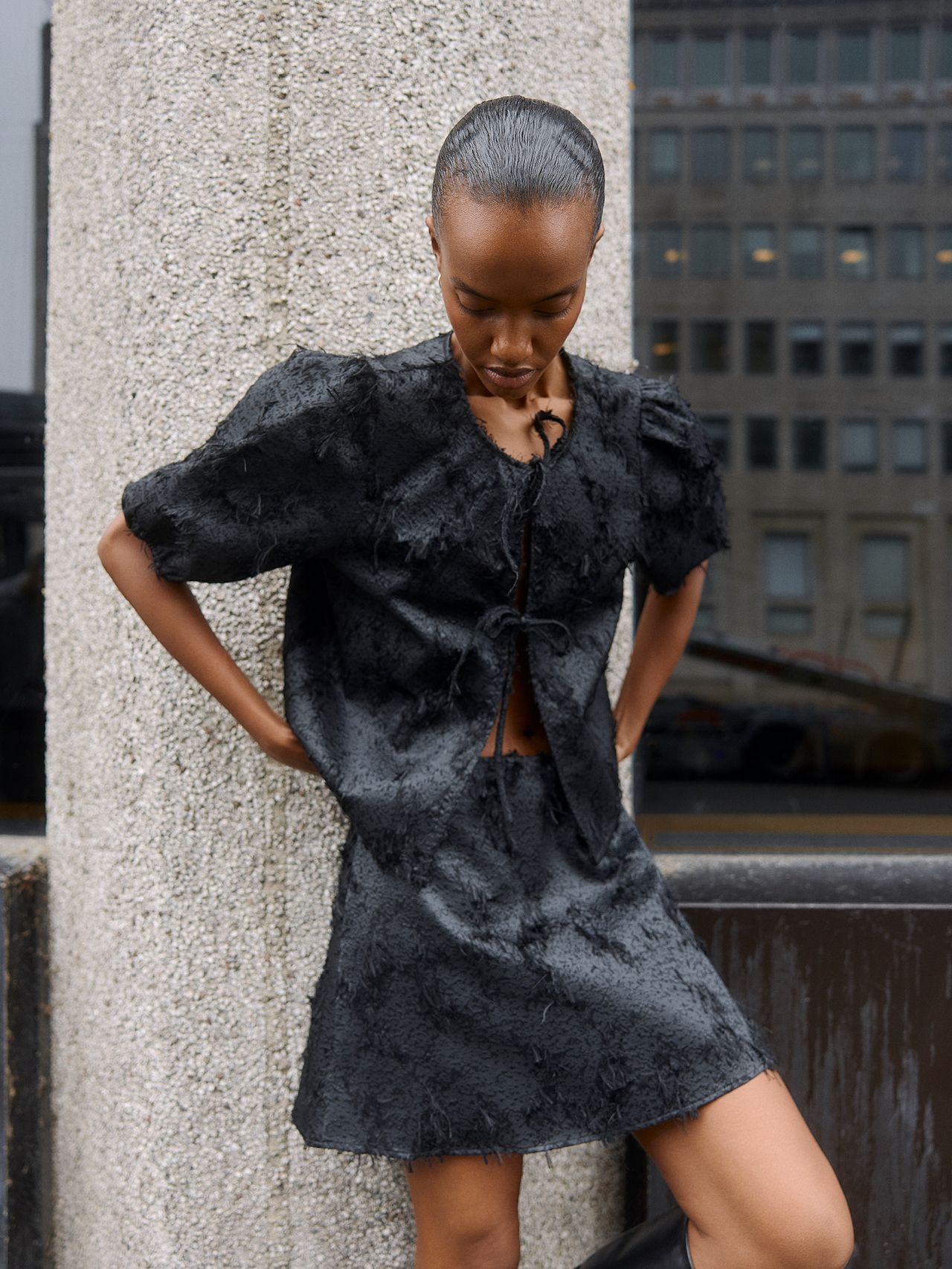 Black woman in a textured two-piece outfit posing by a concrete pillar outdoors