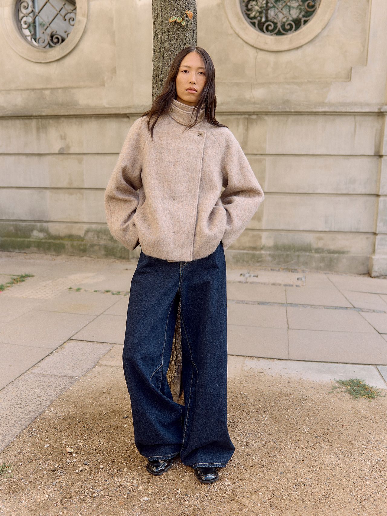 Asian woman in beige wool coat and wide-leg jeans leaning against a tree outdoors