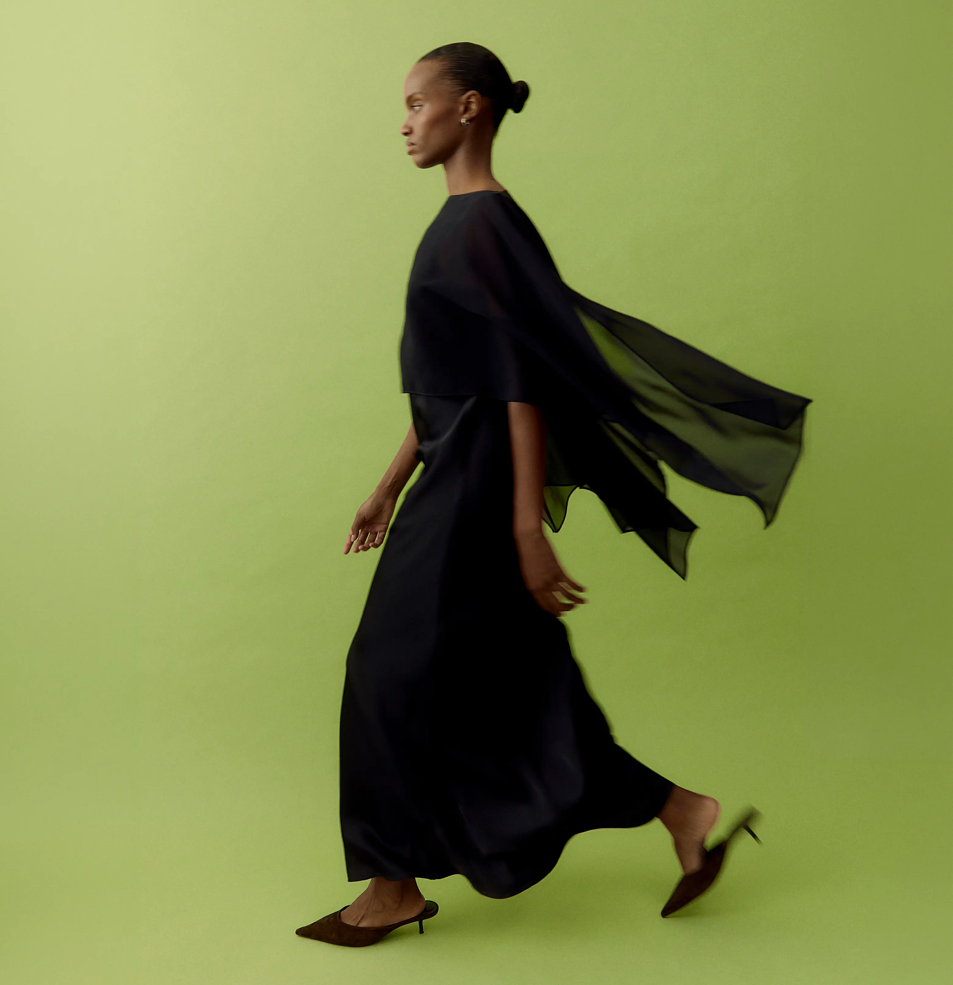 Side view of a woman in a flowing black cape dress and brown mules on a lime green background