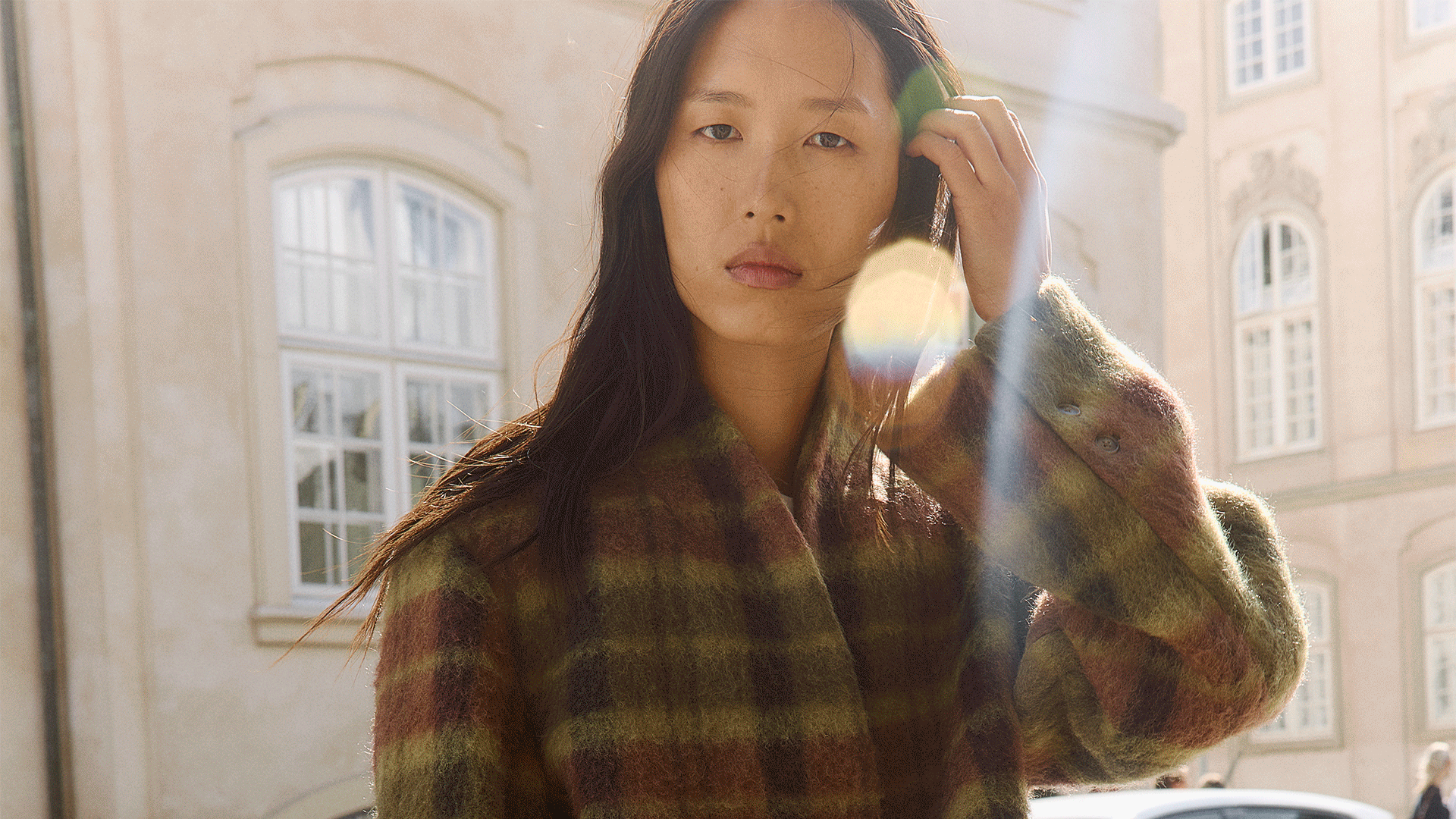 Woman with long dark hair in a plaid wool coat on a sunlit city street.