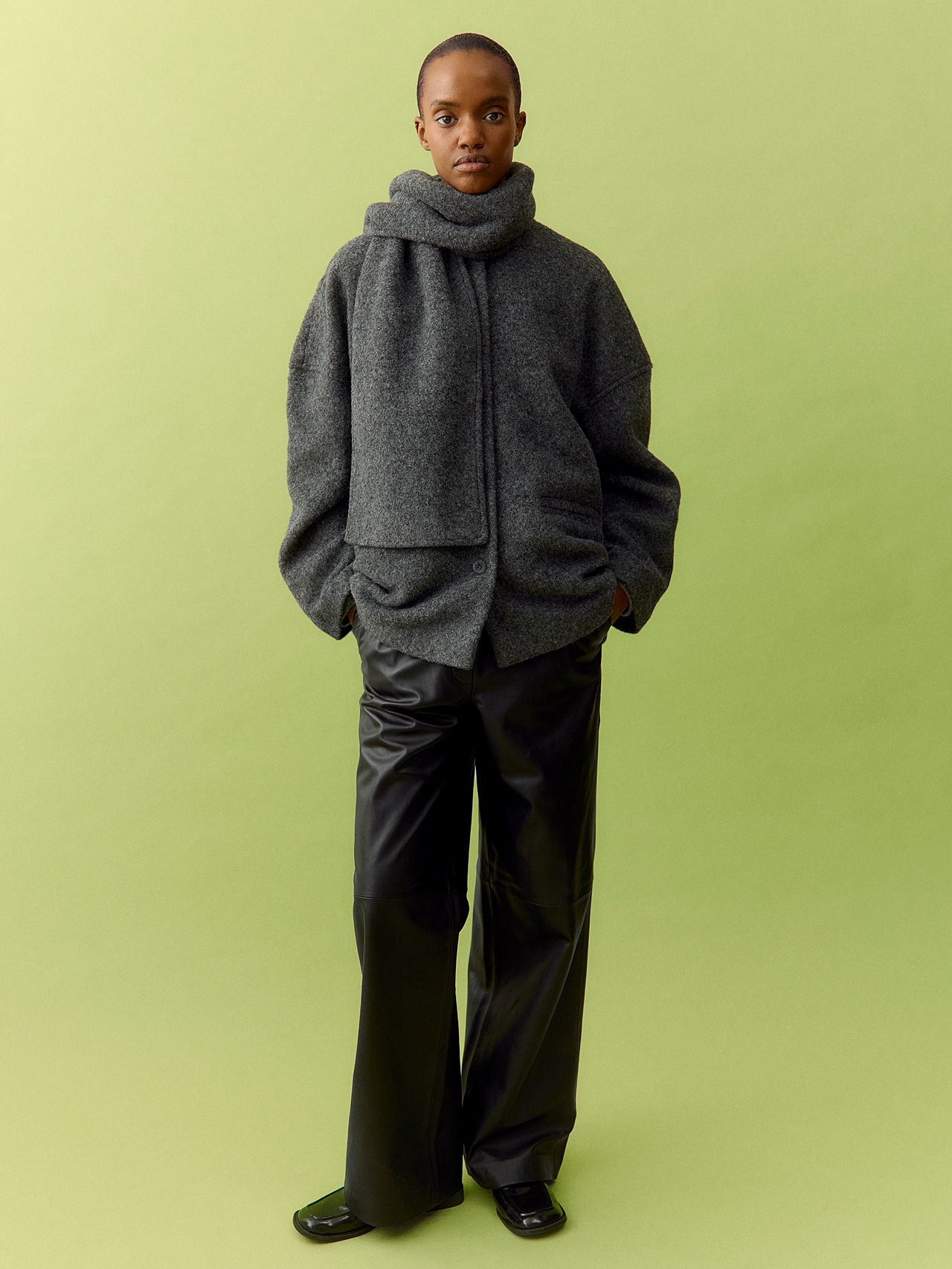 Woman in gray oversized scarf jacket, black wide-leg pants and loafers on lime green backdrop