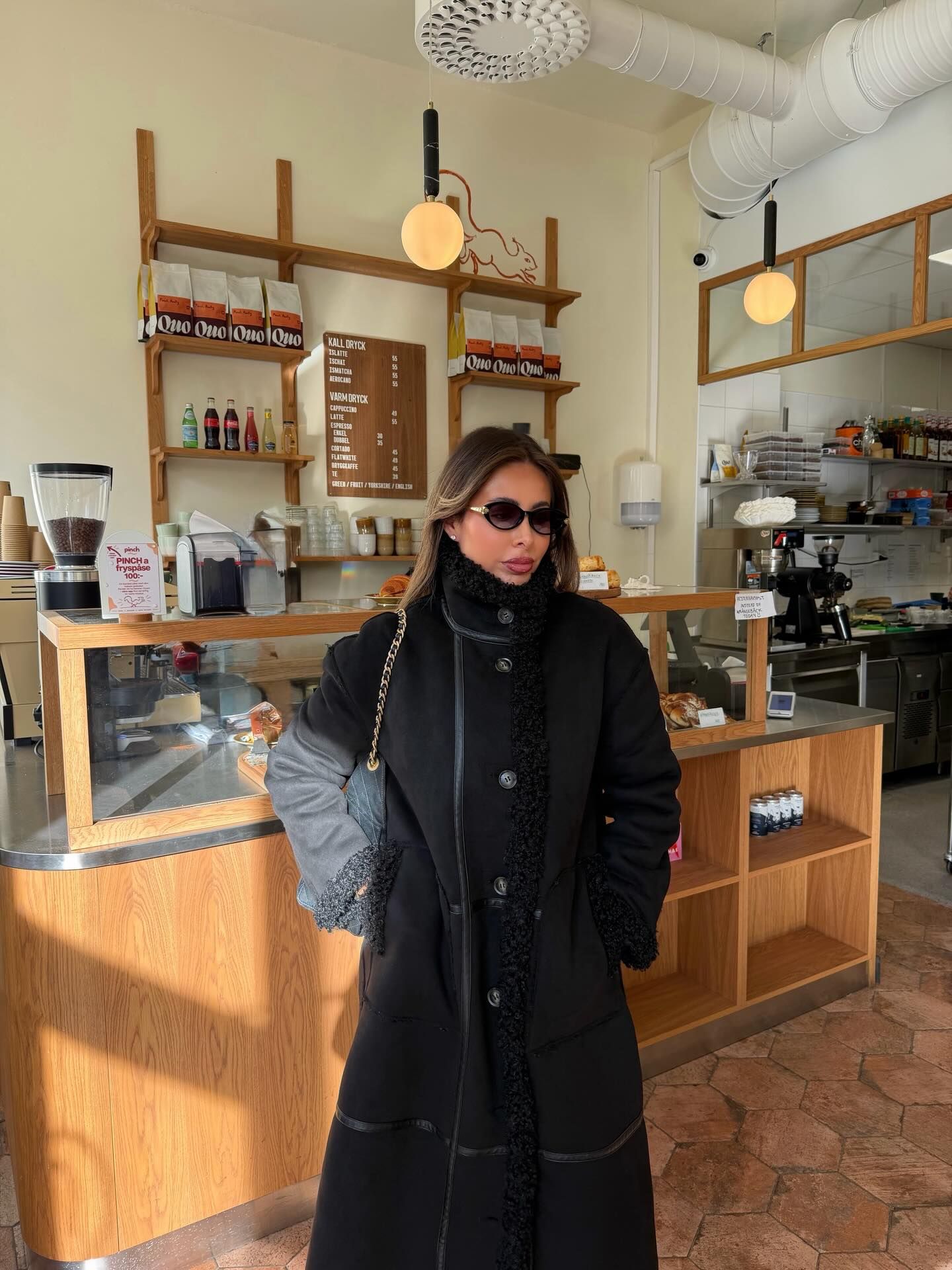 Stilvolle Frau im schwarzen Mantel und Sonnenbrille in einem Café mit Holzregalen