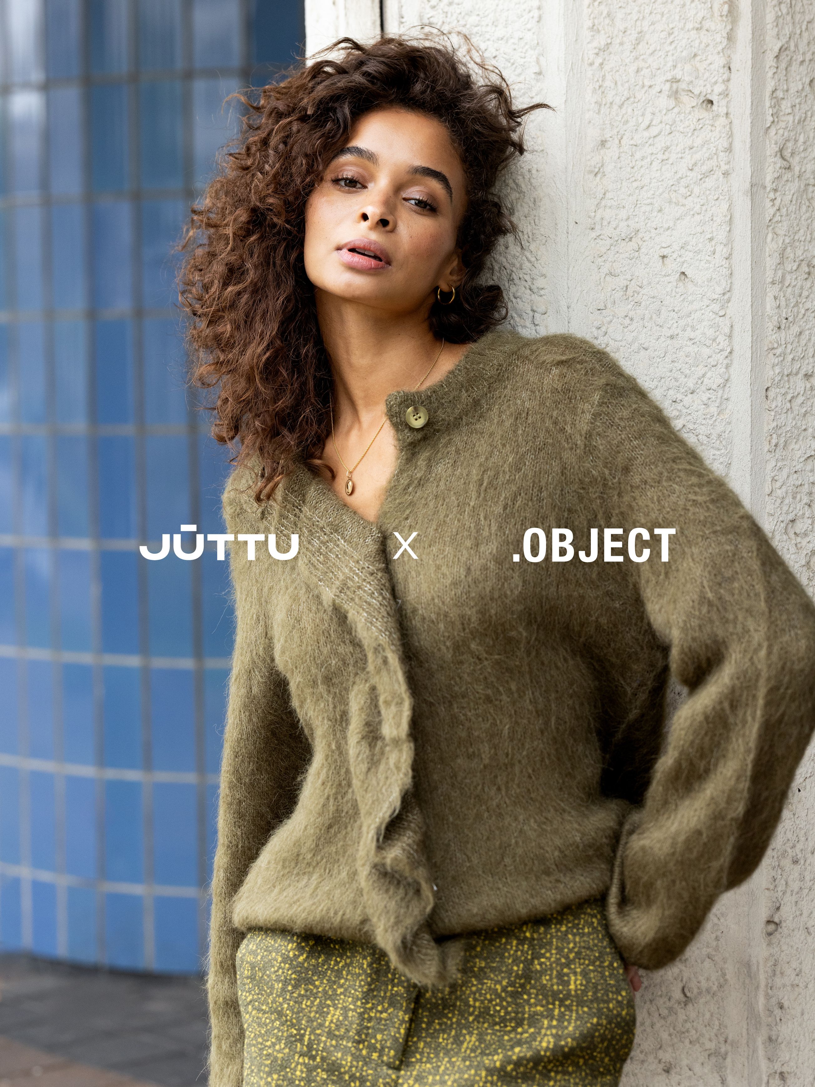 Model met krullen in olijfgroen vest en groene broek leunt tegen blauwe tegelwand.