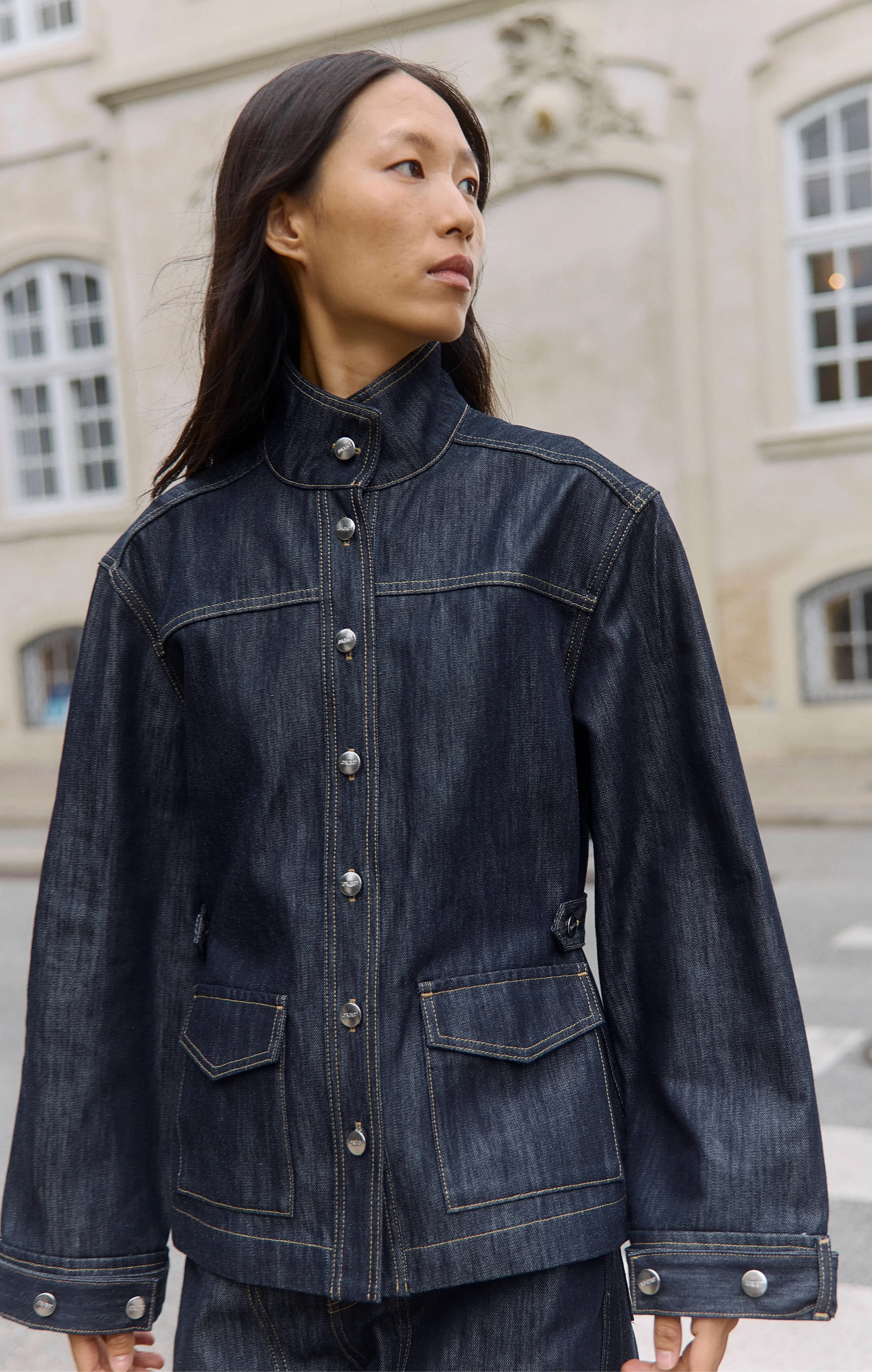 Asian woman in a dark denim jacket, shown in profile against an urban building.