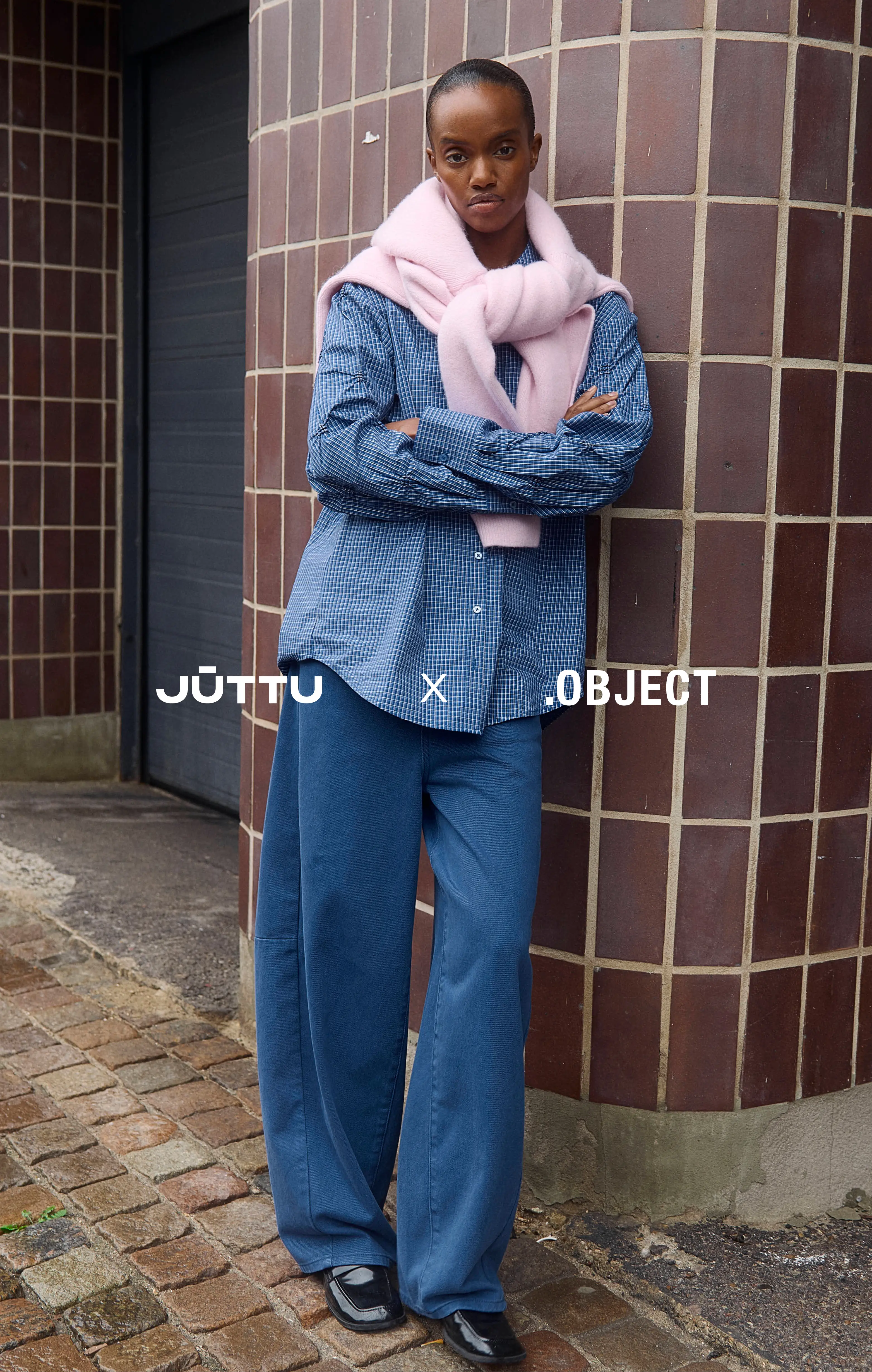 Dark-skinned woman in blue checkered shirt and wide-leg pants, pink sweater over shoulders
