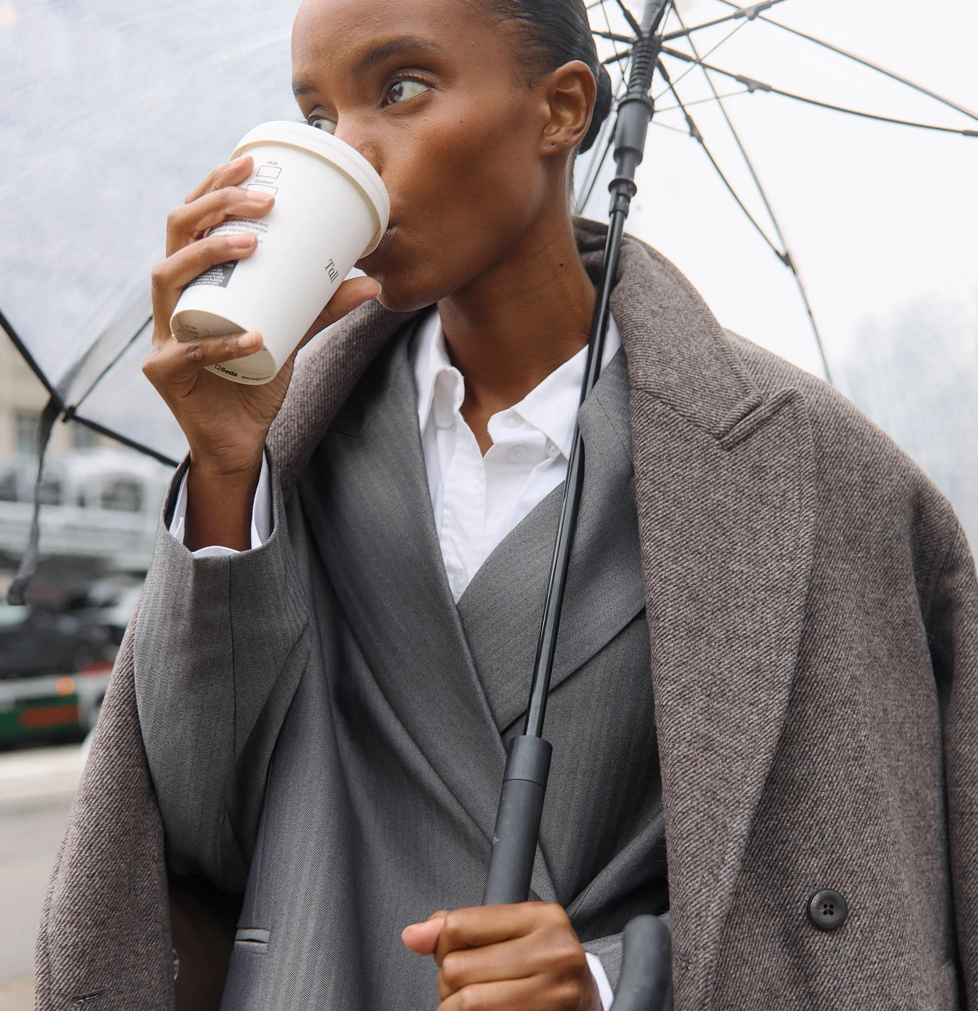 Chic woman in an overcoat drinking coffee under an umbrella on a rainy city street