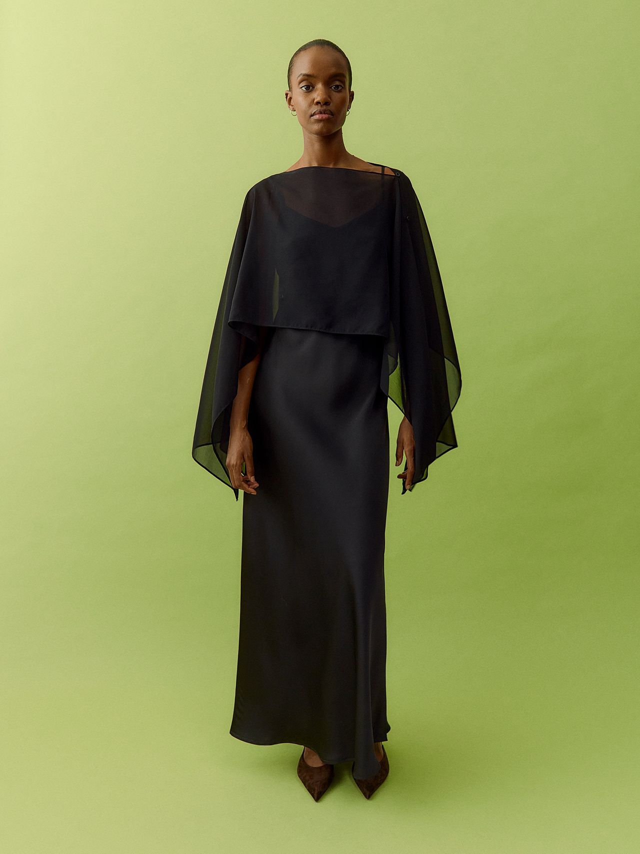Woman in black maxi slip dress with sheer chiffon cape and heels on green background