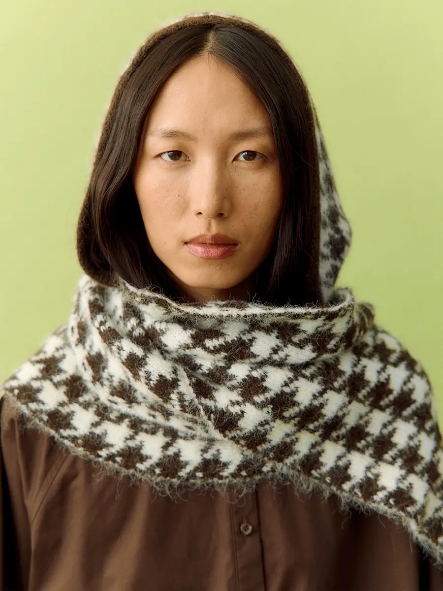 East Asian woman in a brown-and-white knitted hood and scarf against a green backdrop
