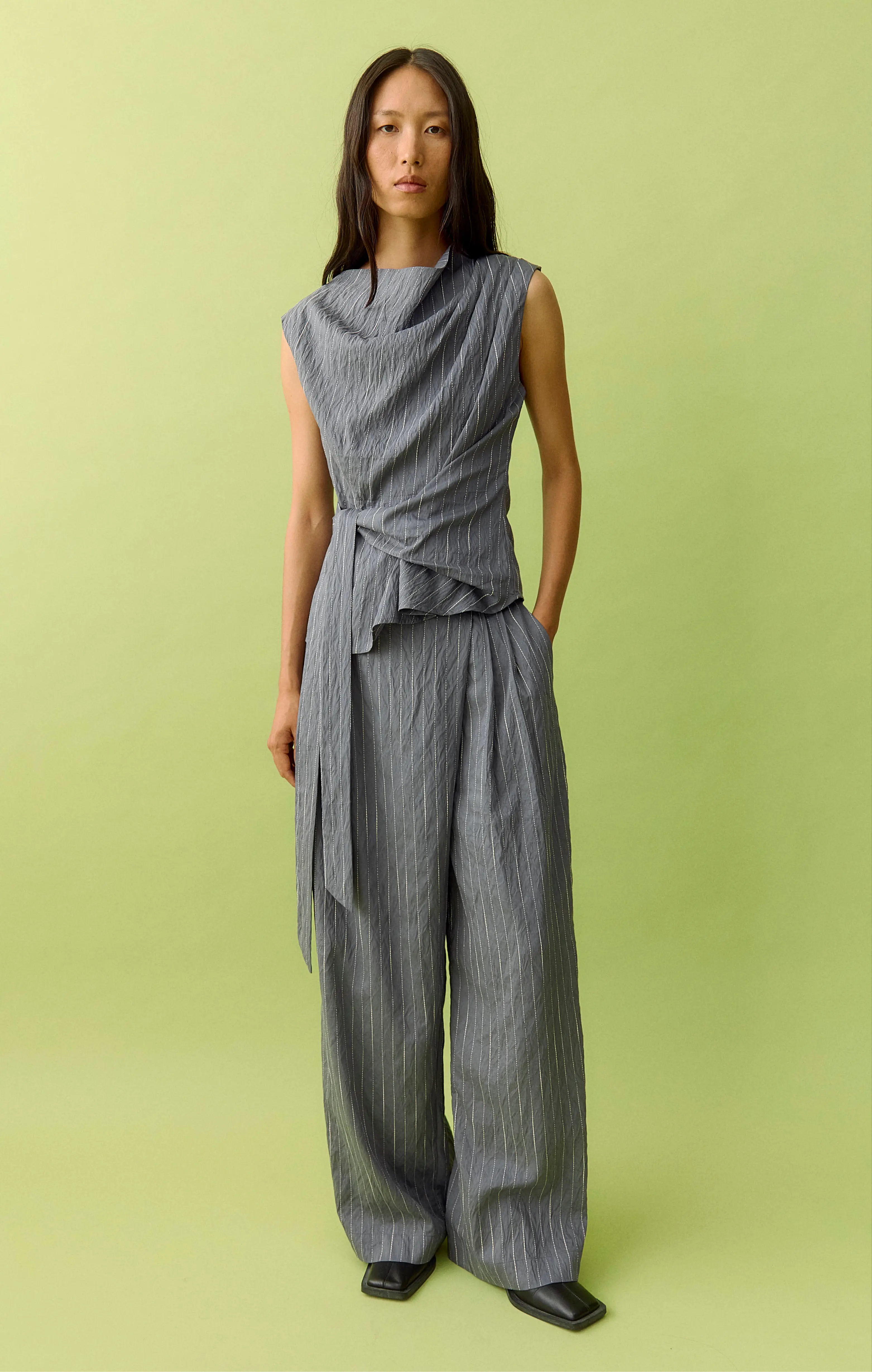 Woman in gray pinstripe draped top and wide-leg pants on green background